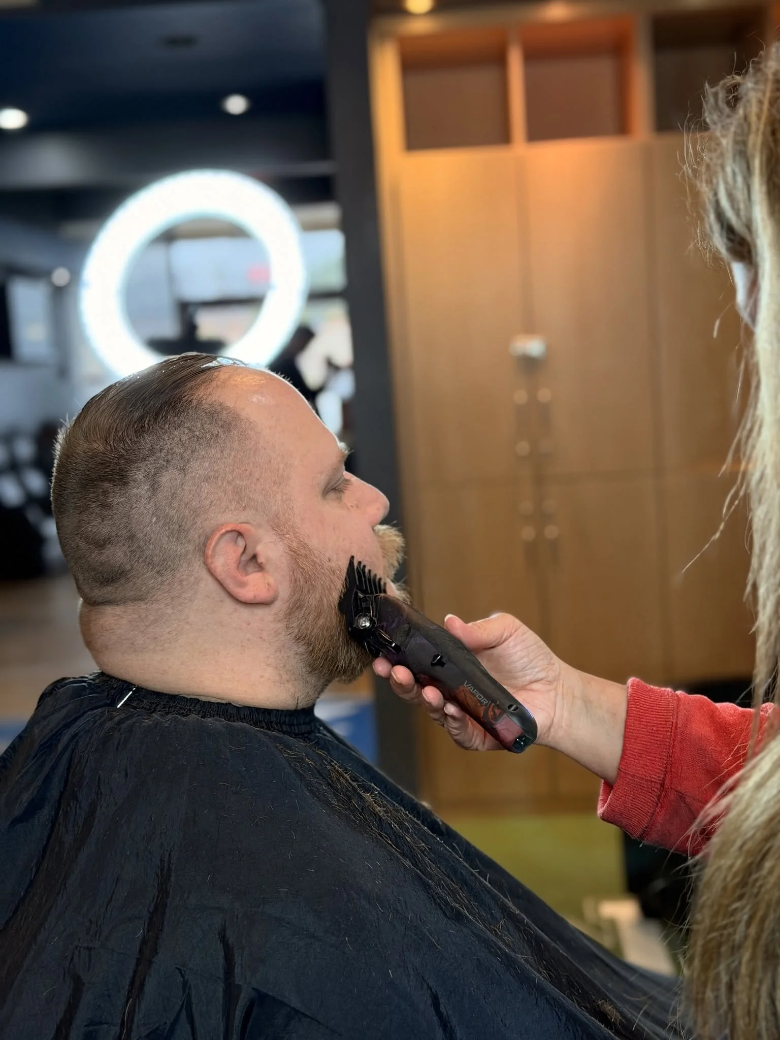 A man getting a haircut at a salon, with a hairdresser using electric clippers on his hair, and a ring-shaped mirror with lights in the background.