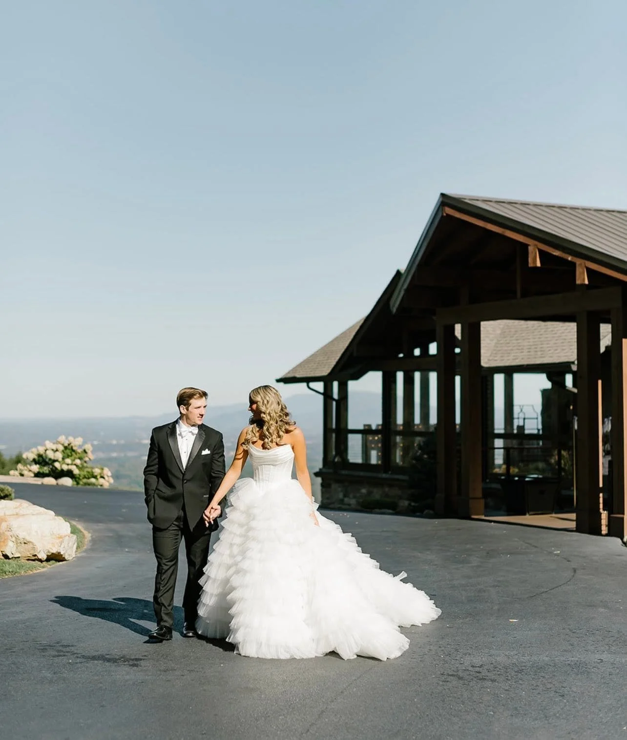 A bride and groom walking hand in hand outside near a wooden pavilion, with mountains and blue sky in the background.