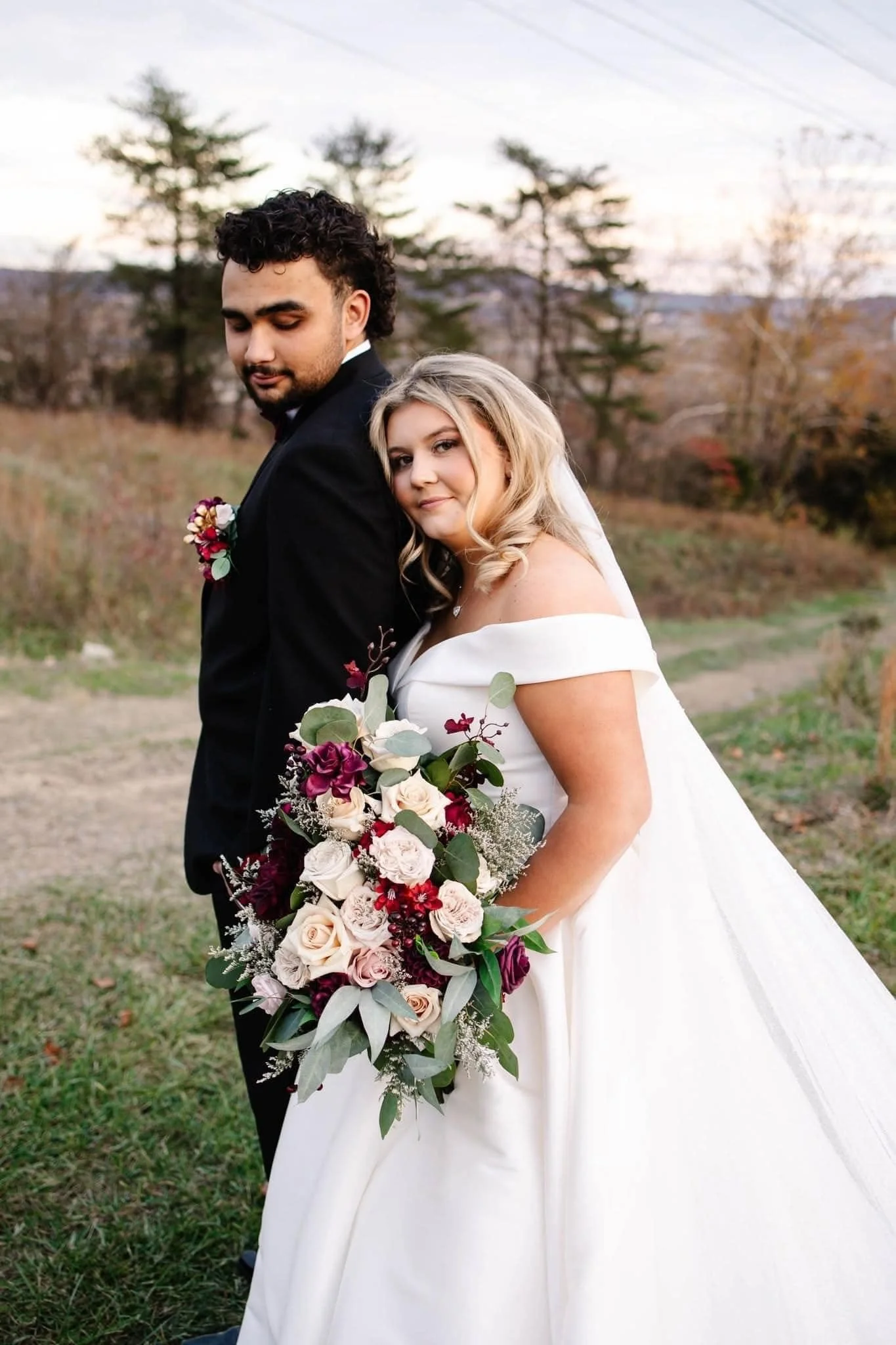 A bride and groom standing outdoors during a wedding photo shoot. The bride is holding a large bouquet of flowers including roses and greenery. The groom is wearing a black suit with a boutonniere. The bride has blonde hair and is wearing an off-shoulder white wedding dress. The background features trees and a partly cloudy sky.