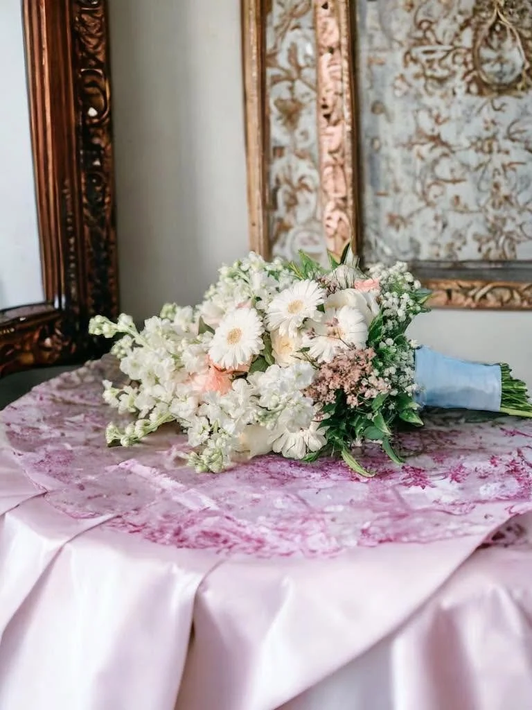 A bouquet of white flowers with pink accents laying on a pink tablecloth in front of a mirror and ornate wallpaper.