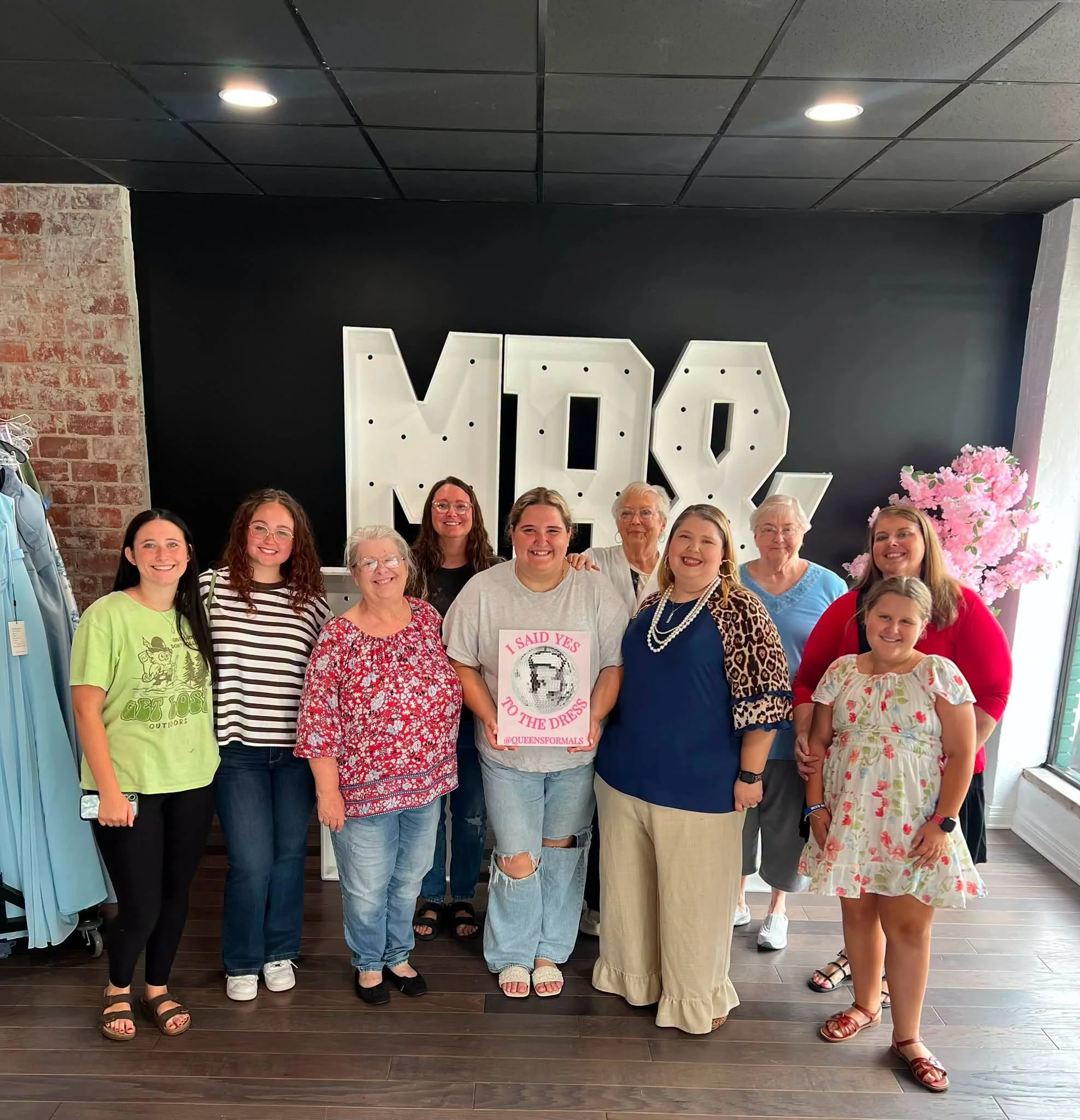 Group of diverse women standing together in front of large decorative letters and flowers, smiling for a photo, inside a room with dark walls and a brick column.