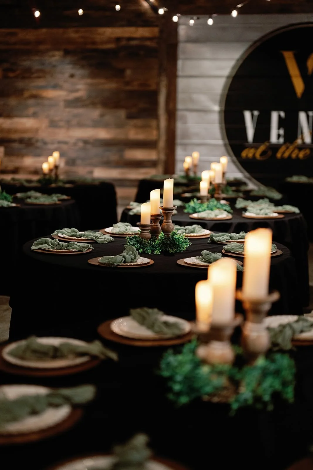 Rustic indoor dining setup with candles and green napkins on black tablecloths, wooden walls, and a large circular sign with partially visible letters.