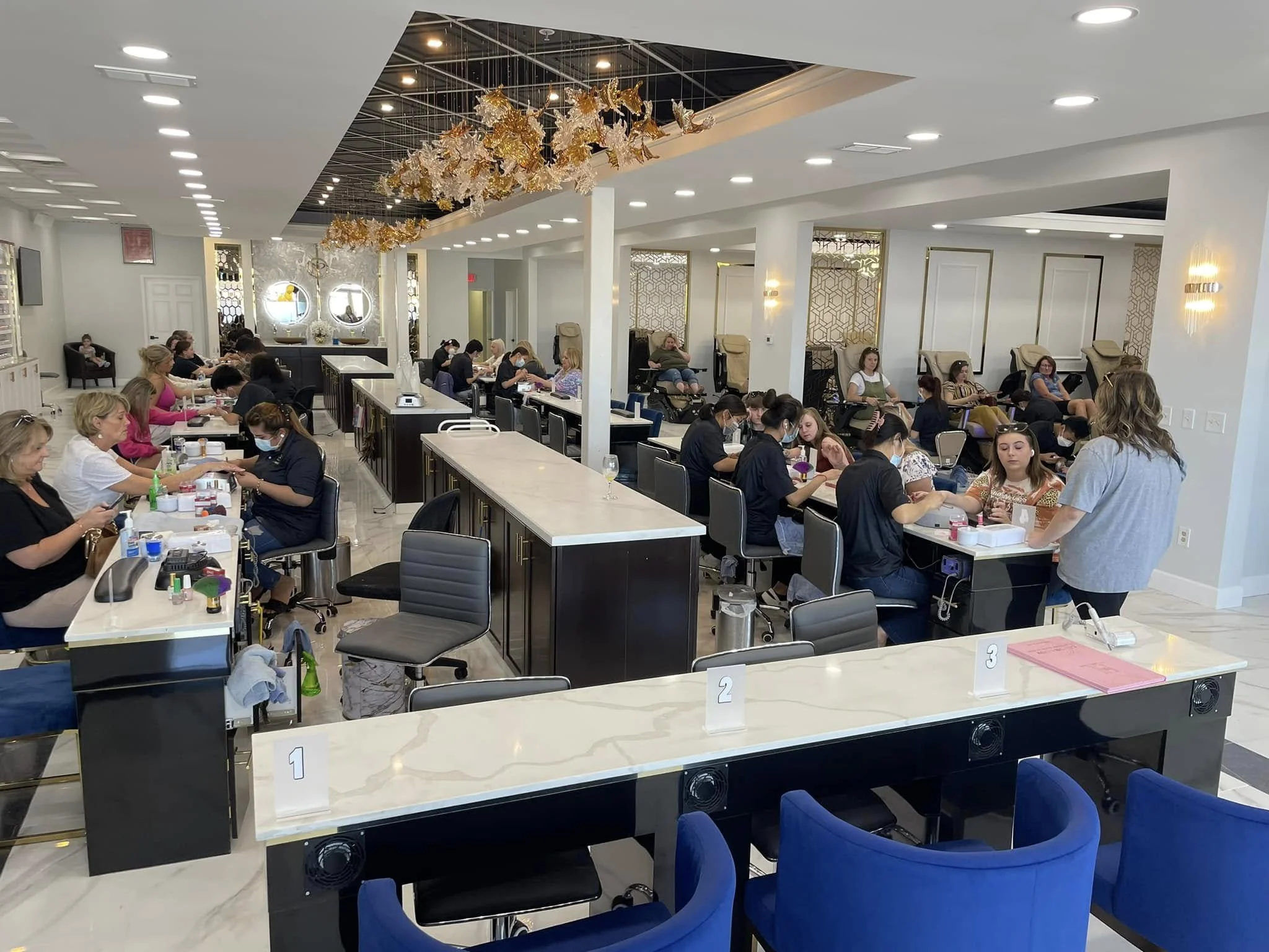 A nail salon with multiple women getting manicures, some sitting in massage chairs. The salon has modern decor with decorative lighting, gold accents, and a unique ceiling installation with hanging metallic and glass art pieces.