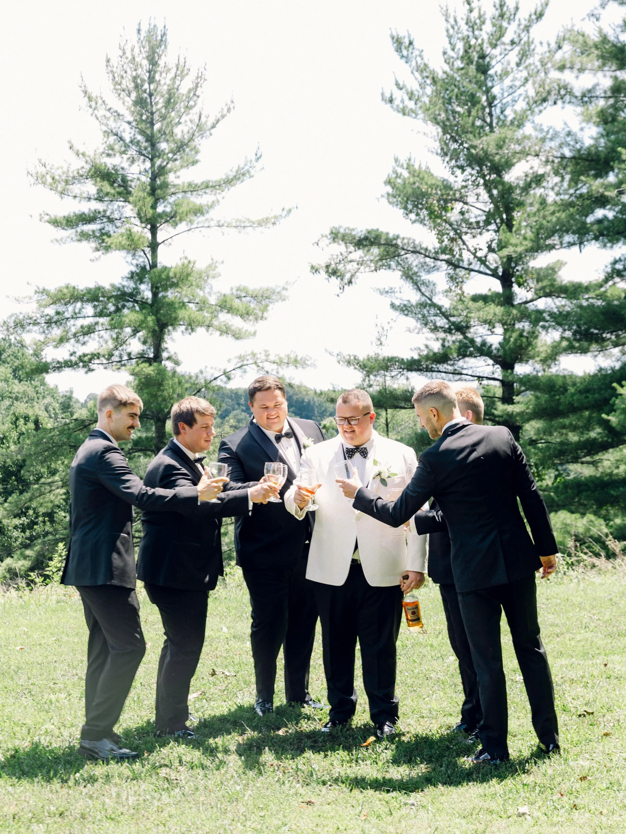 A group of men in tuxedos and a man in a white tuxedo jacket with a black bow tie, standing outdoors on grass with trees in the background, holding glasses and a bottle, socializing.