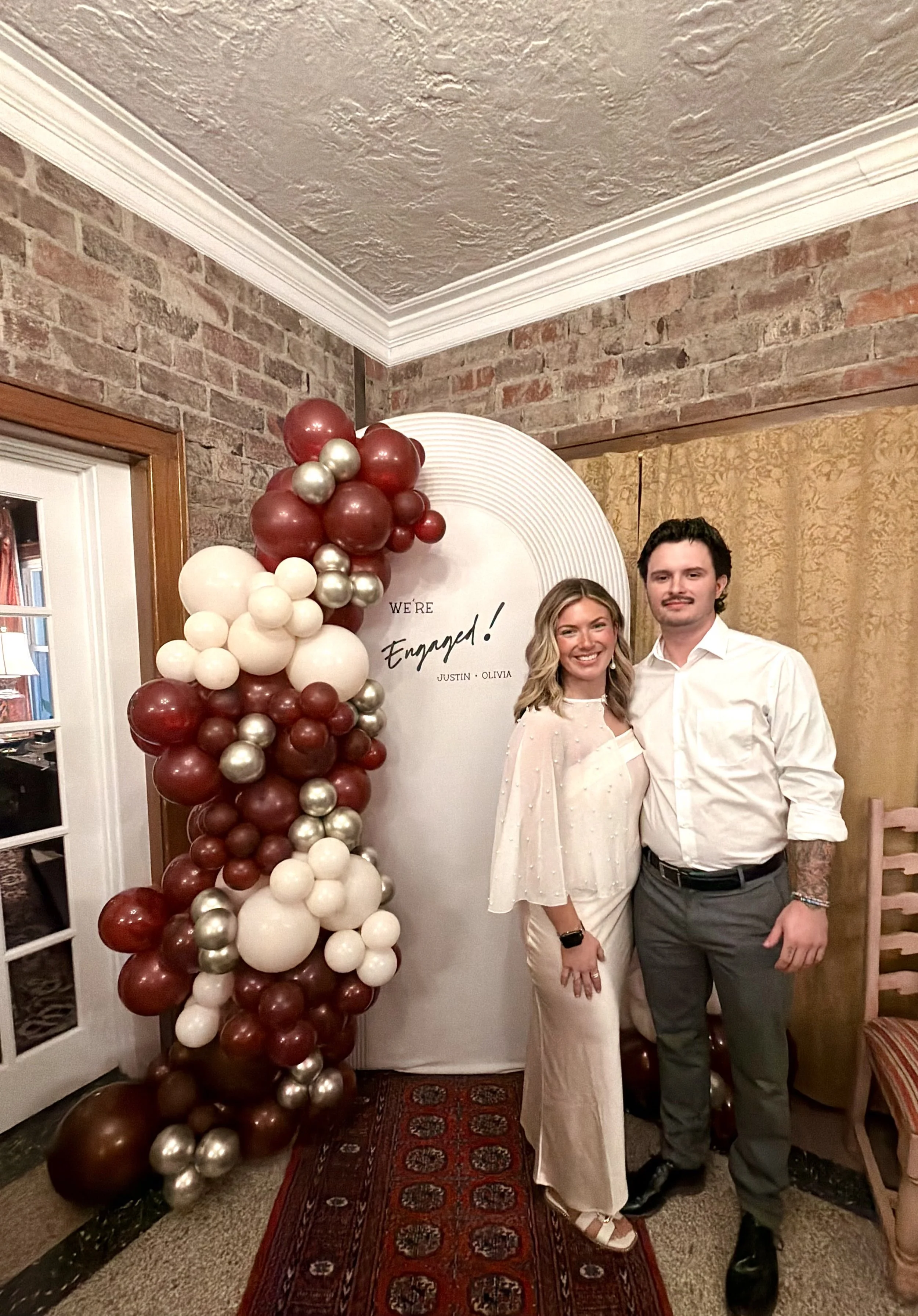 A smiling woman and a man stand together in front of a balloon decoration at a celebration. The woman wears a white dress and the man a white shirt and gray trousers.