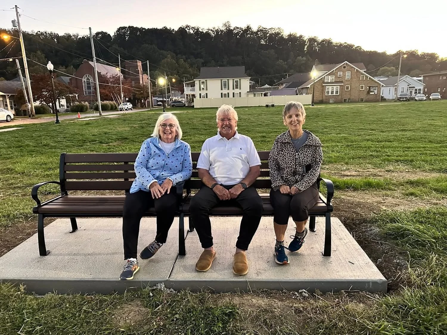Three elderly adults sitting on a park bench with a grassy field and houses in the background during sunset.