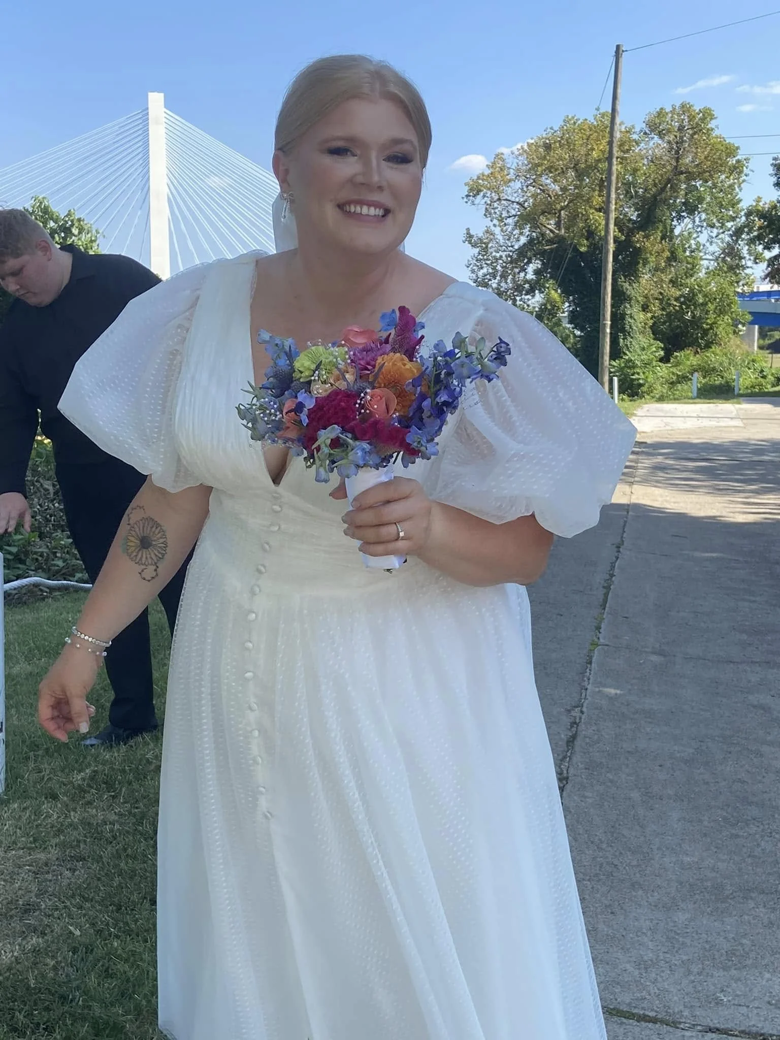 A woman in a white dress holding a colorful bouquet of flowers, standing outdoors with a bridge and trees in the background.