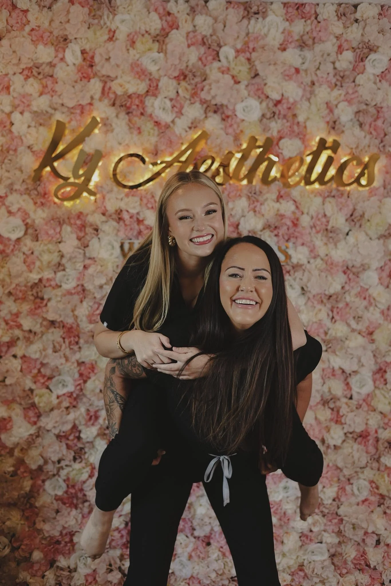 Two women smiling and hugging in front of a floral pink and white flower wall with gold lettering that reads 'Ky Aesthetics'.