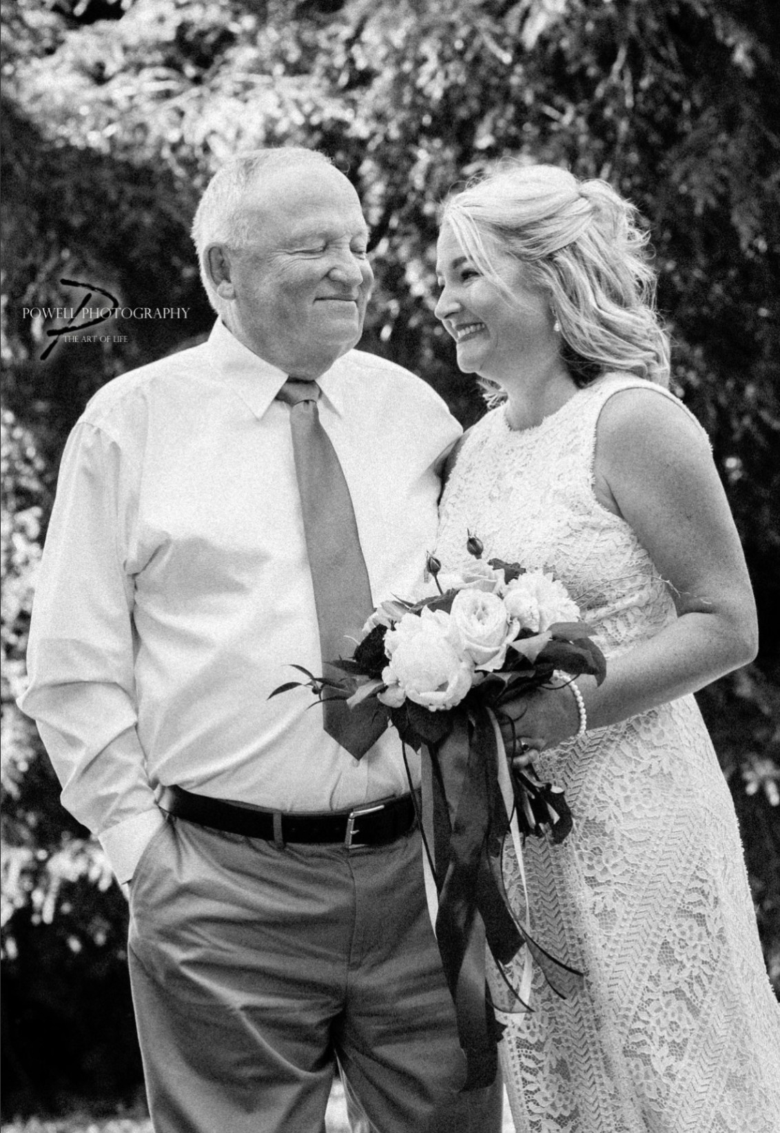 A man and woman, both smiling, stand close together outdoors. The woman is holding a bouquet of flowers. The man wears a dress shirt and tie, and the woman wears a lace dress.