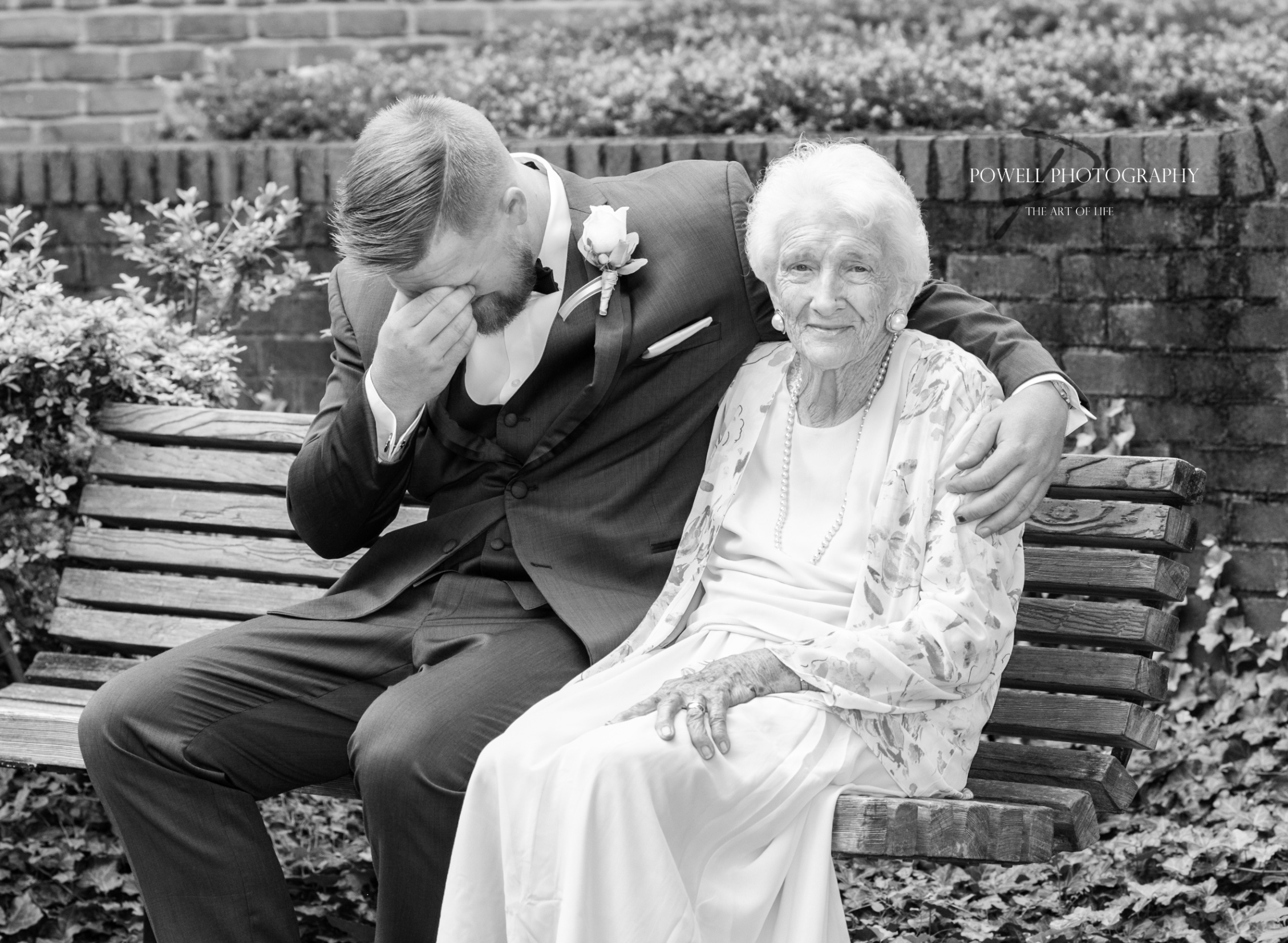 A man in a suit and an elderly woman sitting on a wooden bench outdoors, with the man emotional, wiping his face, and the woman smiling, embracing him. There are flowers and a brick wall in the background.