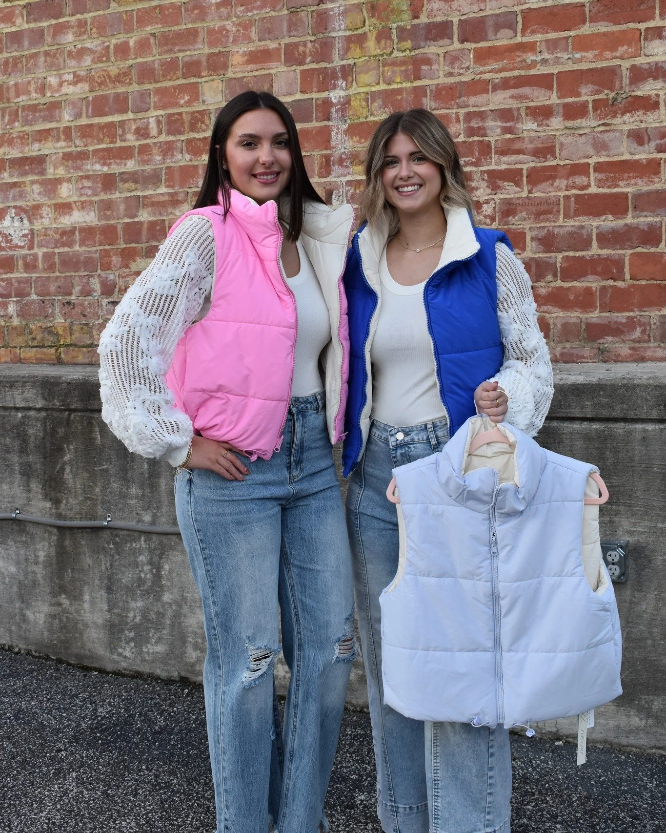 Two young women standing outdoors in front of a brick wall, smiling, with one holding a light-colored vest on a hanger.
