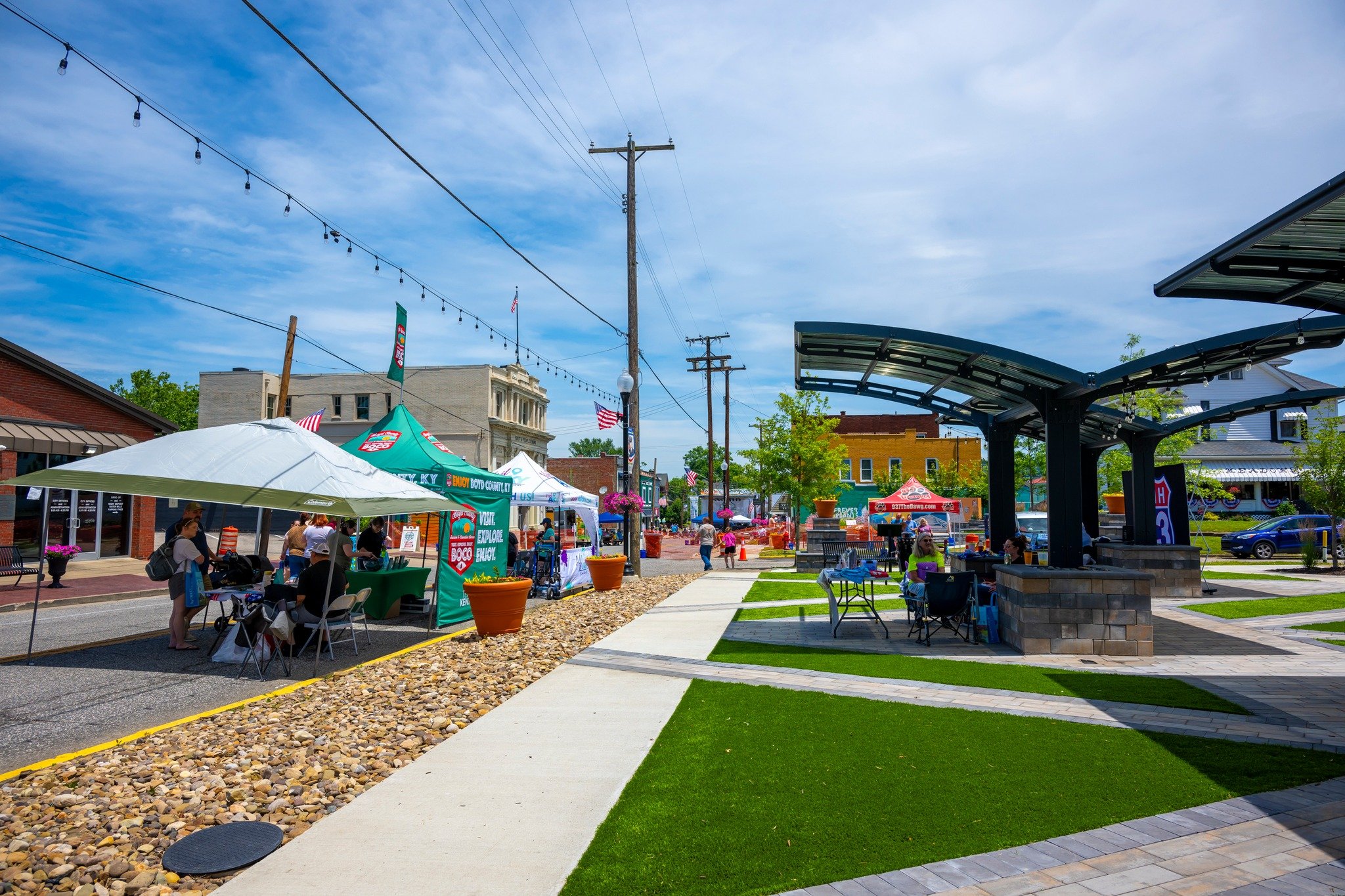 A lively outdoor street fair with tents, booths, and people walking and sitting. There are string lights overhead, potted plants, and a modern pavilion. The sky is mostly clear with some clouds.