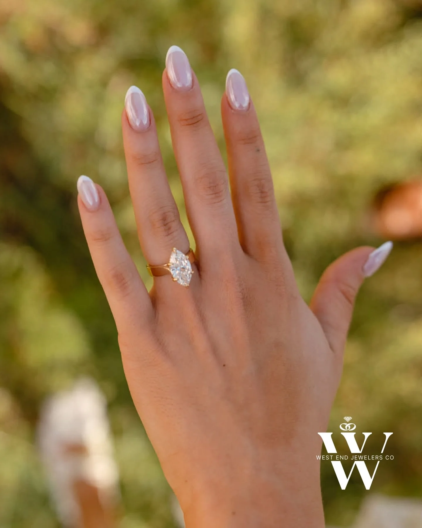 A woman's hand showing a large, marquise-cut diamond engagement ring on her ring finger. The background is blurred greenery with warm sunlight.