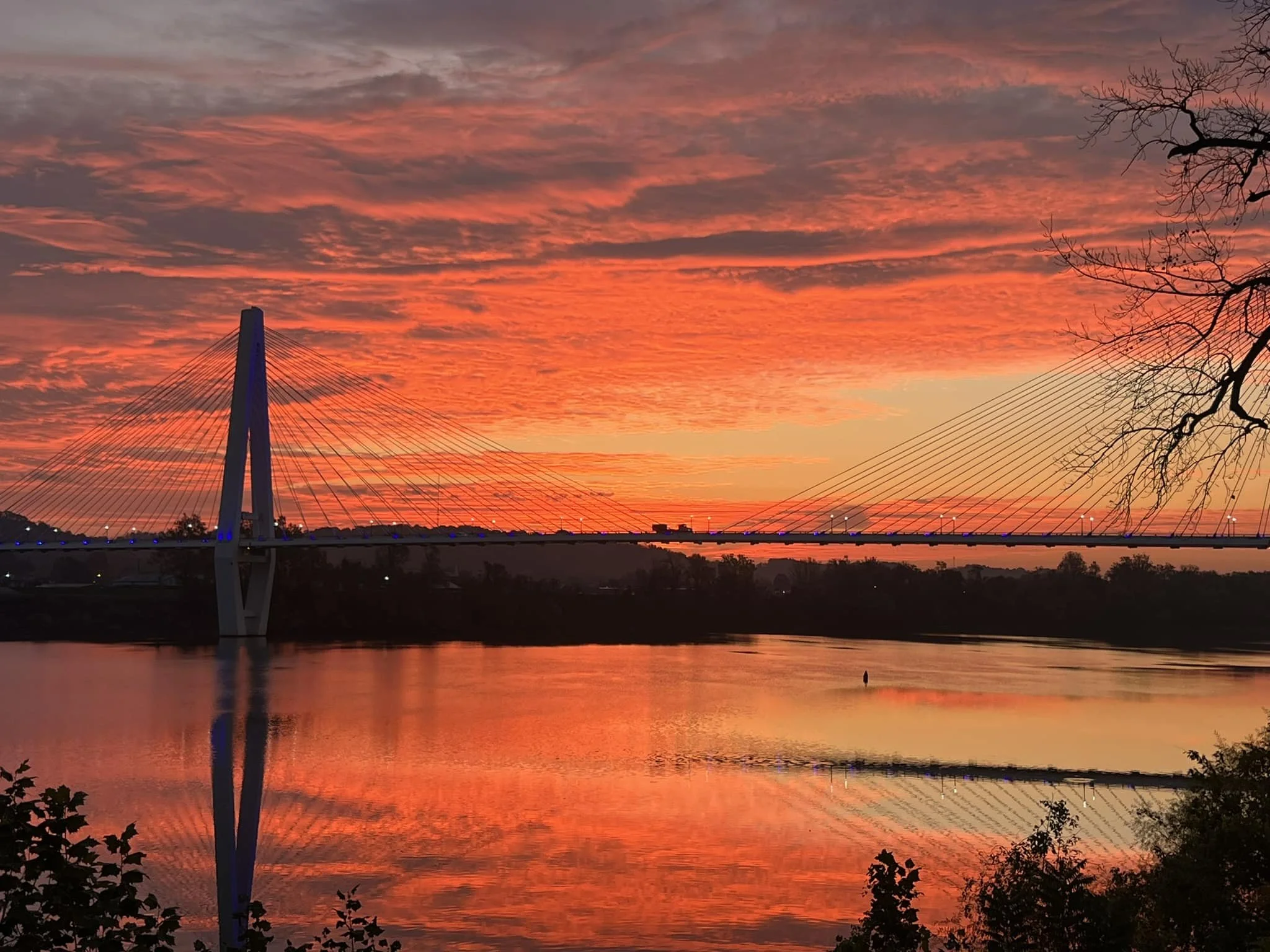 A bridge over a river during sunset with orange and pink clouds in the sky, and reflections of the sky and bridge on the water.