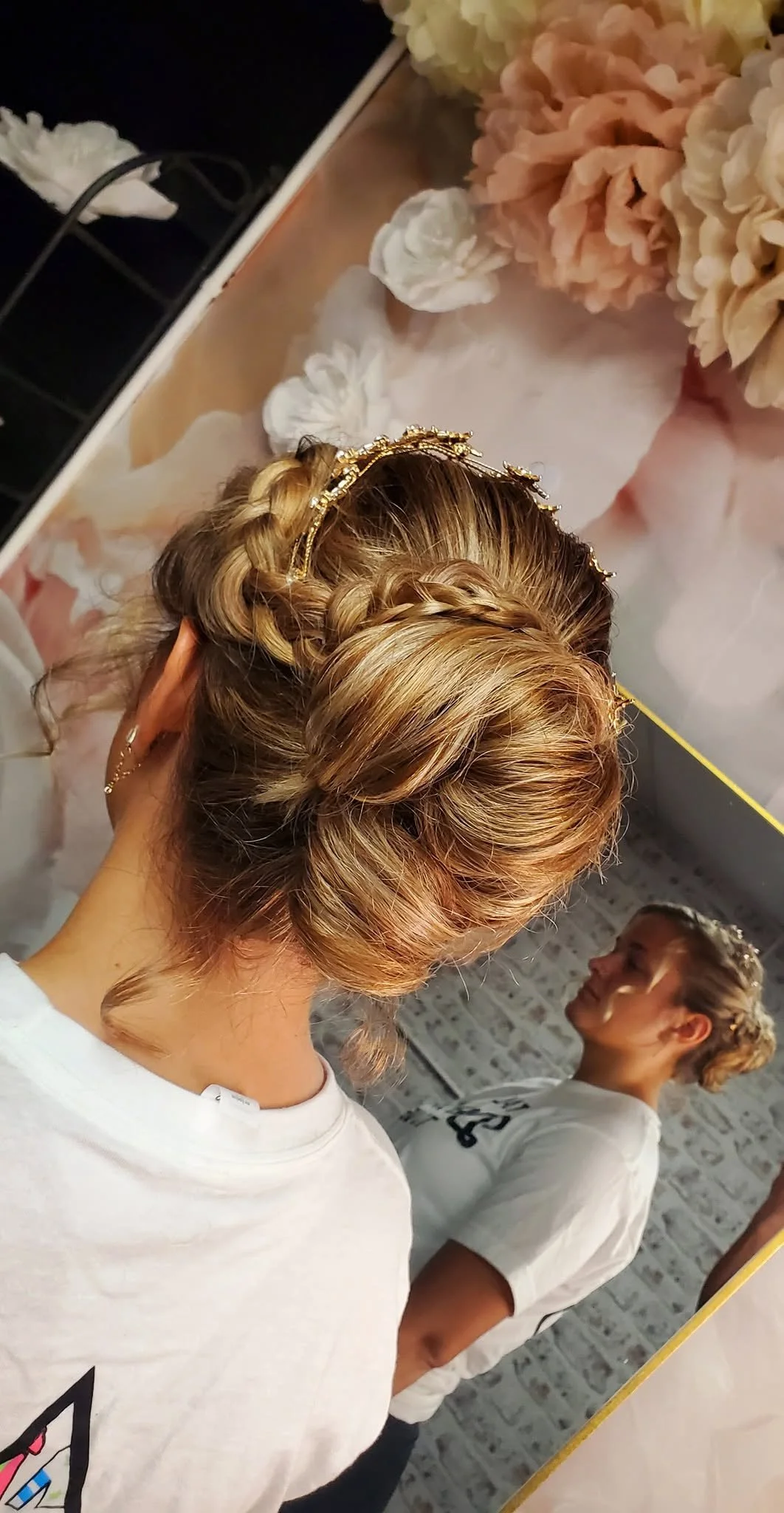 A woman with braided blonde hair and a gold headband seen from behind, looking into a mirror with a young girl also with braided hair and a white T-shirt, in a room decorated with cream and peach flowers and a white brick wall.