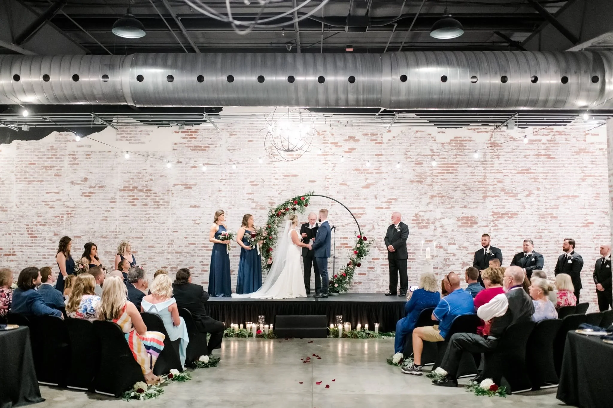 A wedding ceremony with a bride and groom exchanging vows under a floral arch, surrounded by family and friends in a spacious industrial-style venue with a large metal air duct and exposed brick wall.