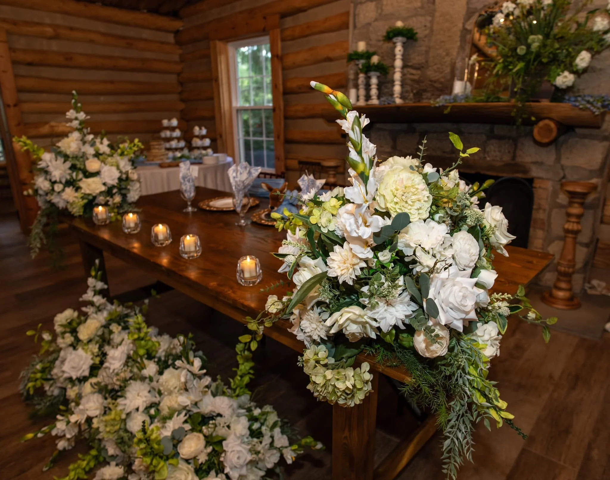 A rustic indoor setting with a wooden table decorated with floral arrangements of white flowers and greenery, surrounded by candles in small holders. The background includes logs, a window, and additional floral decorations.