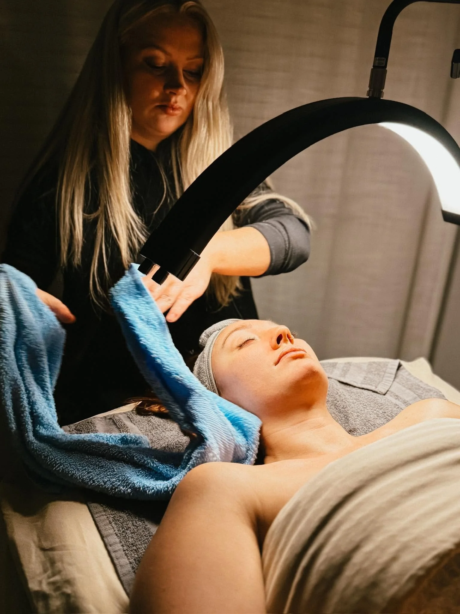 A woman with long blond hair receiving a facial or skincare treatment while lying on a treatment bed, with a towel draped over her chest and a black therapeutic light device directed at her face.