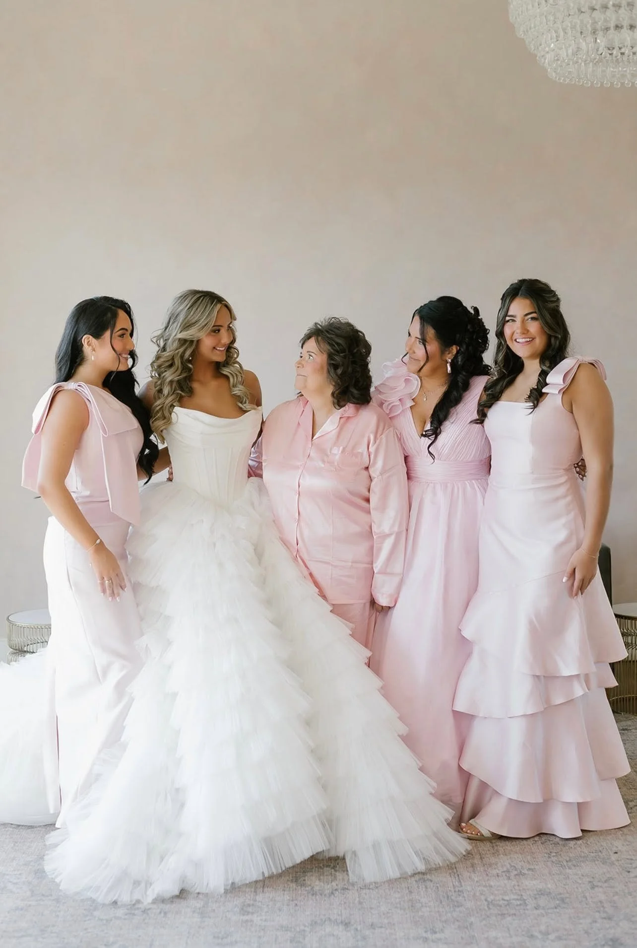 A bride in a white wedding gown surrounded by five women in pink dresses, standing in a room with a chandelier and plain wall.