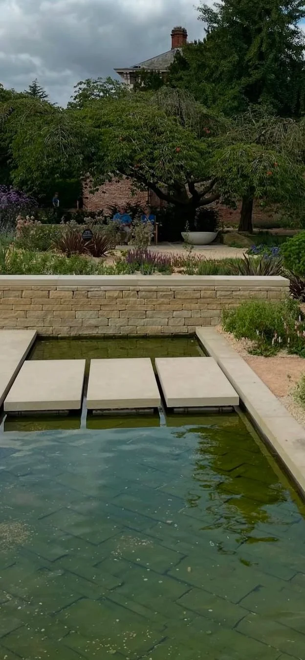 A garden with a pond, stone pathway, lush greenery, and a house with a brick wall in the background.