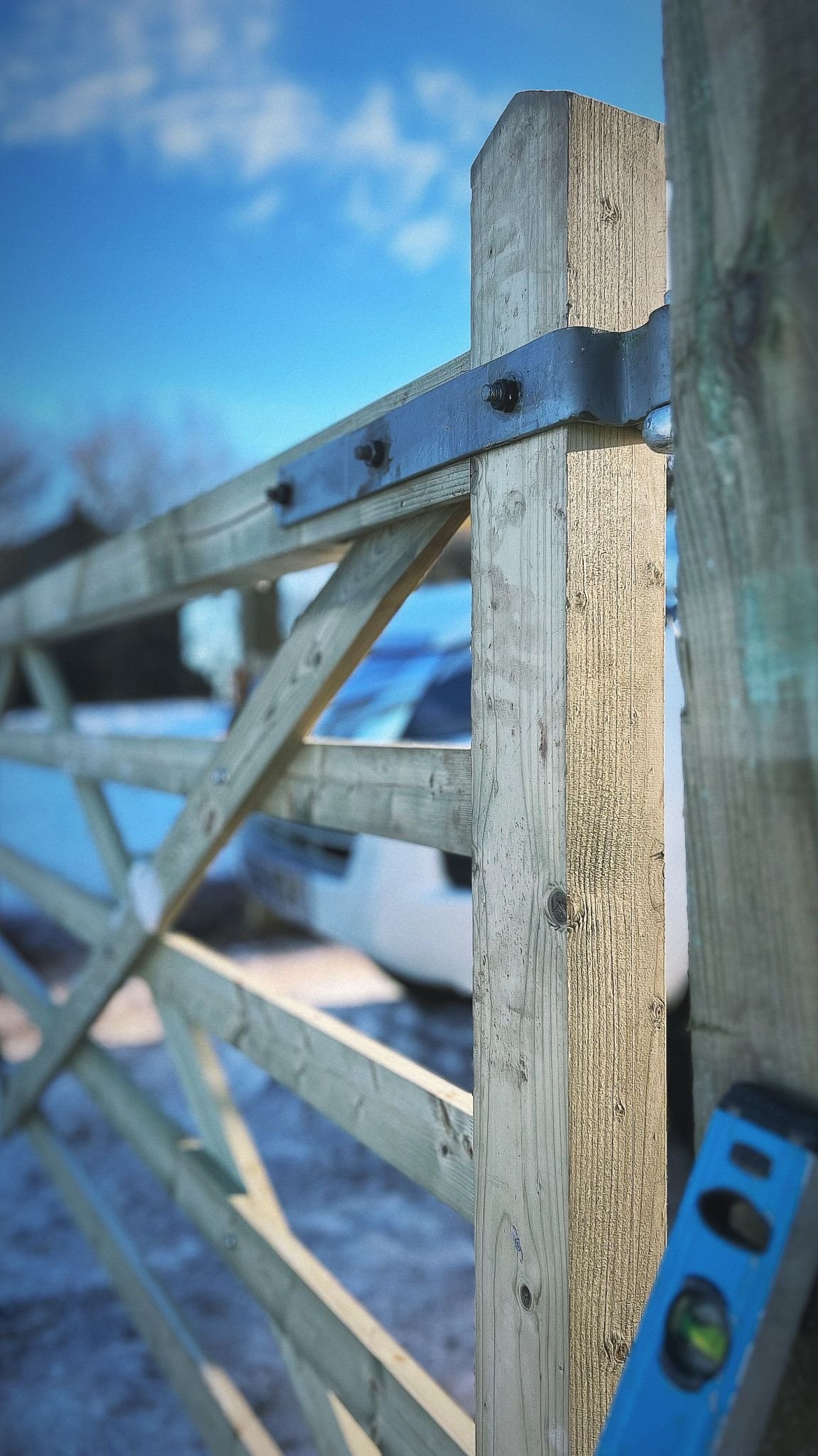 Close-up of a wooden fence or railing under construction with a blue spirit level resting on it. The background shows a parking lot and a cloudy sky.