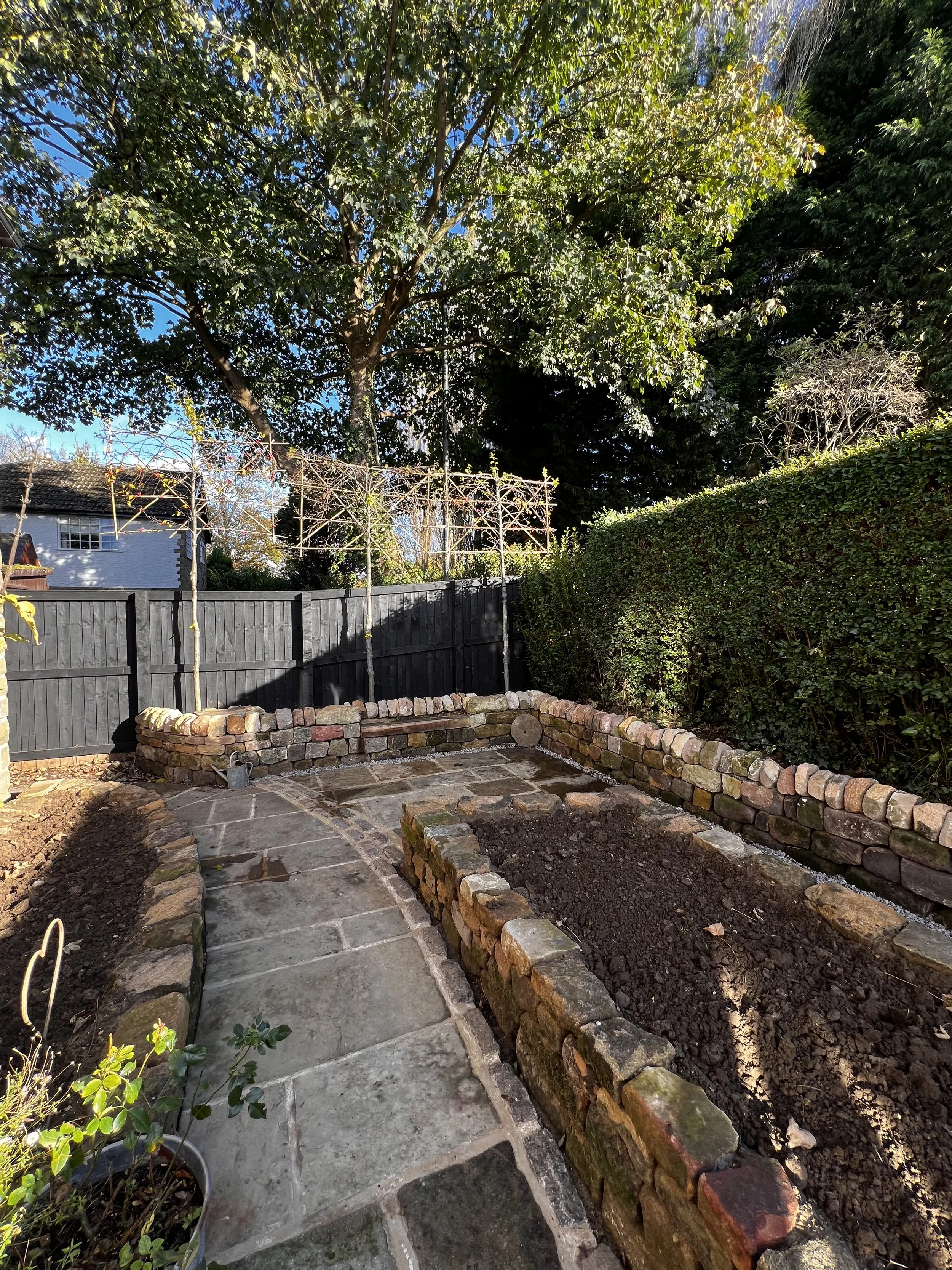 Garden yard under construction with stone pathway, brick raised beds, and a black wooden fence, surrounded by trees and shrubs.