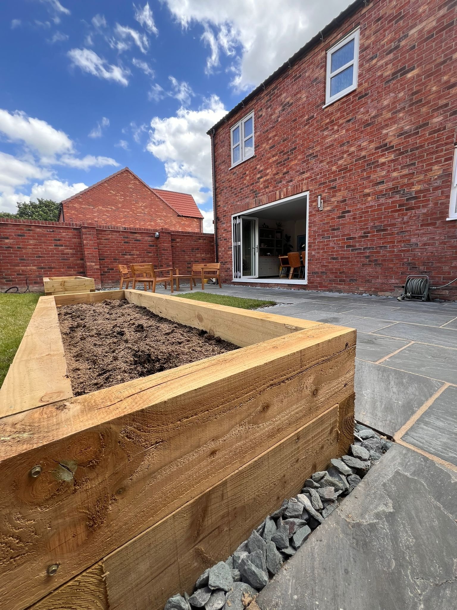 A backyard patio with a wooden raised garden bed filled with soil, outdoor furniture including a table and chairs, and a brick house with open sliding glass doors leading to an indoor space, under a partly cloudy sky.