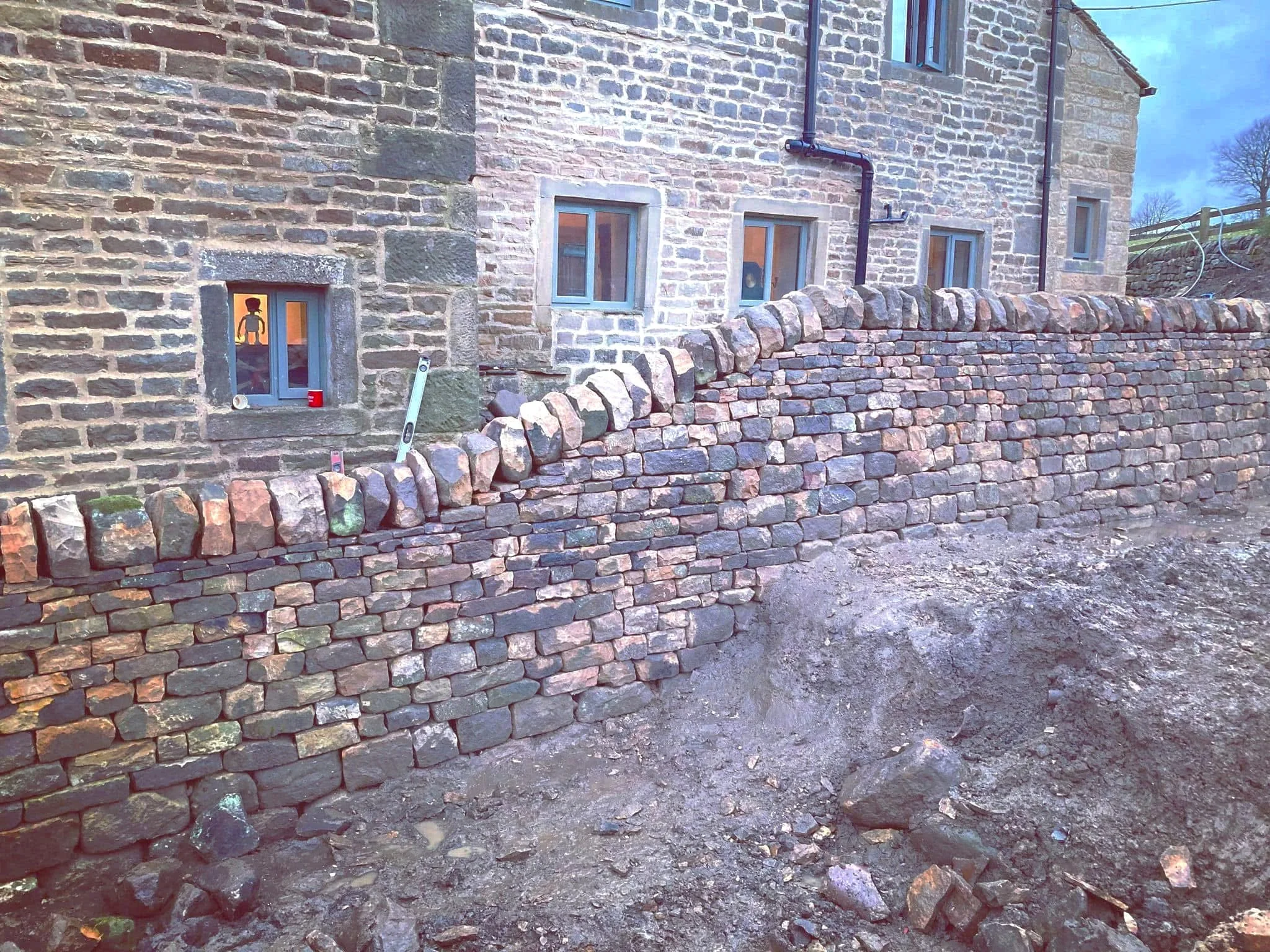 A dry stone wall under construction in front of a brick house with three windows, with a level tool and a red cup placed on the wall, and an uneven dirt ground in the foreground.