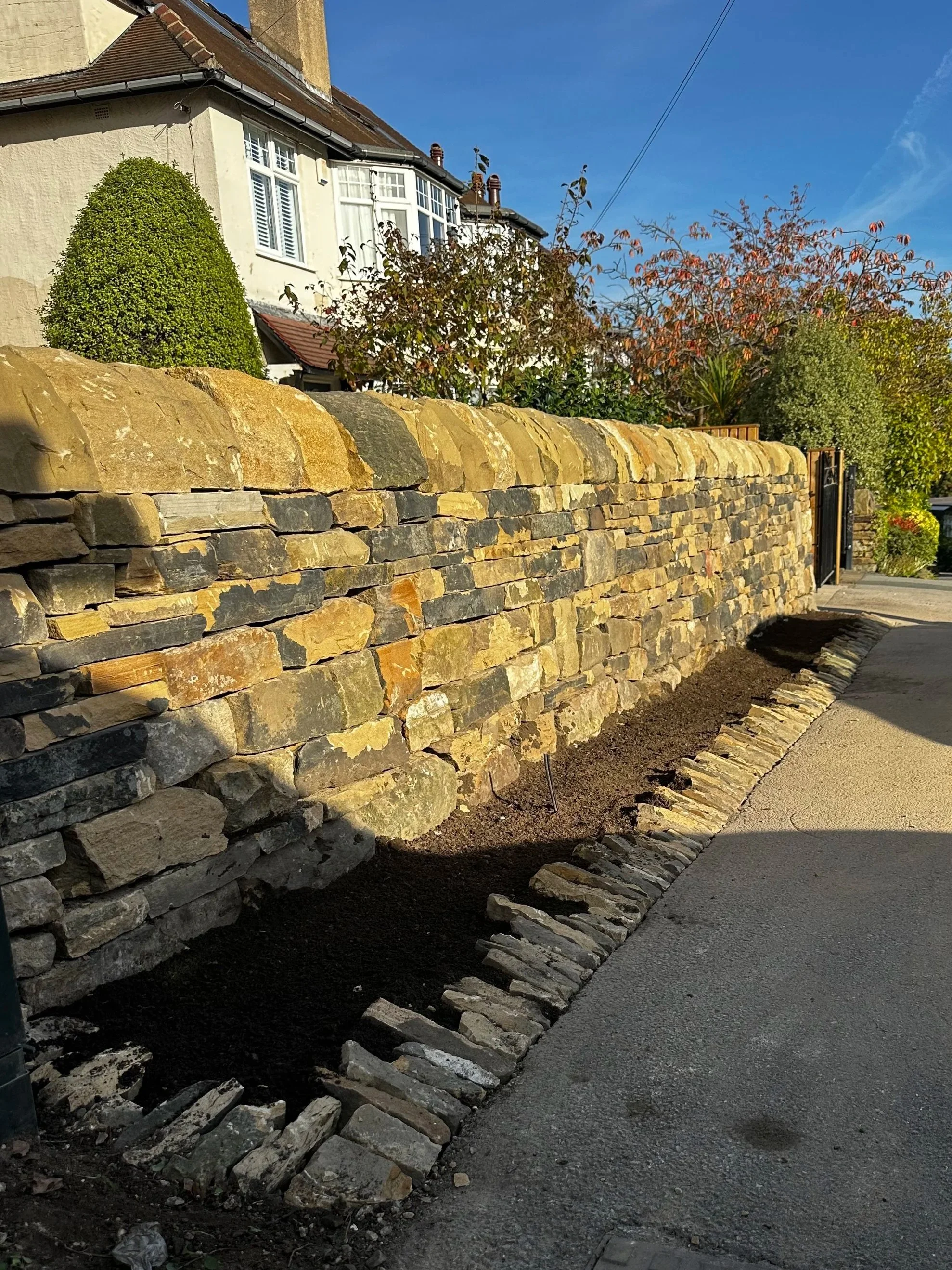 A dry stone wall along a sidewalk with soil in front, under a clear blue sky.
