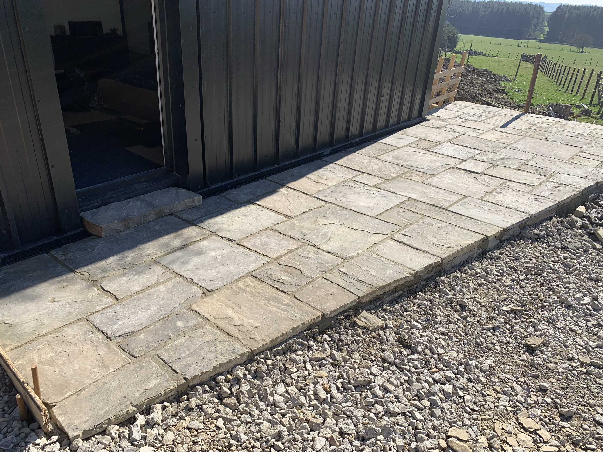 A newly constructed stone patio outside a building with a black metal exterior wall, overlooking a green field and distant trees.