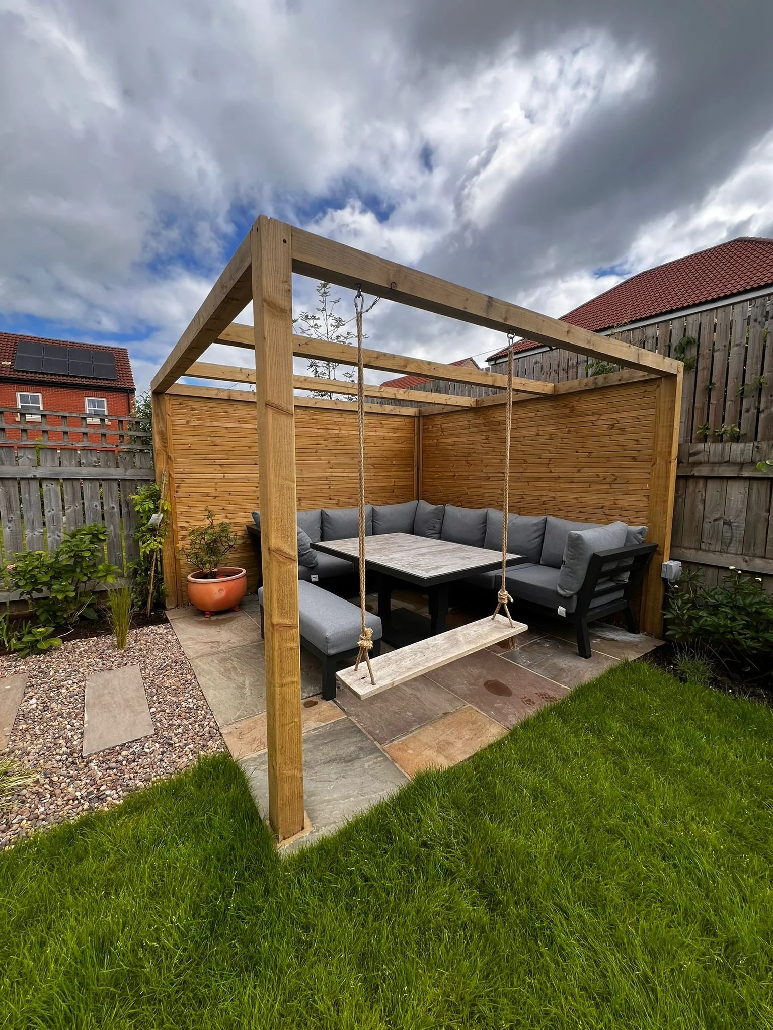 Backyard seating area with a wooden pergola, gray cushioned bench, a table, and a swing, surrounded by a wooden fence and green grass, under cloudy sky.