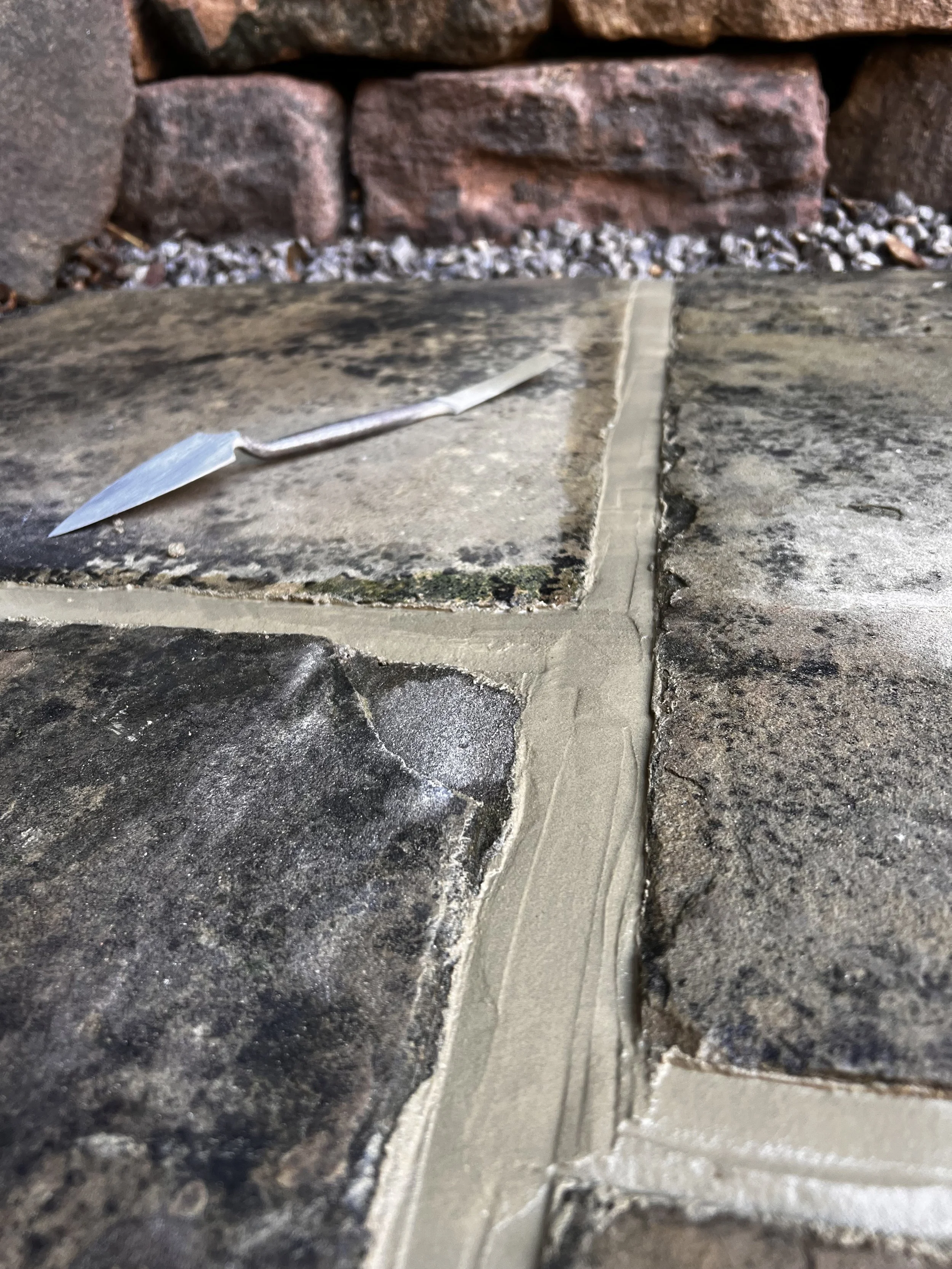Close-up of a stone patio with newly applied mortar between the tiles, with a small trowel tool placed on the patio, and a brick wall in the background.