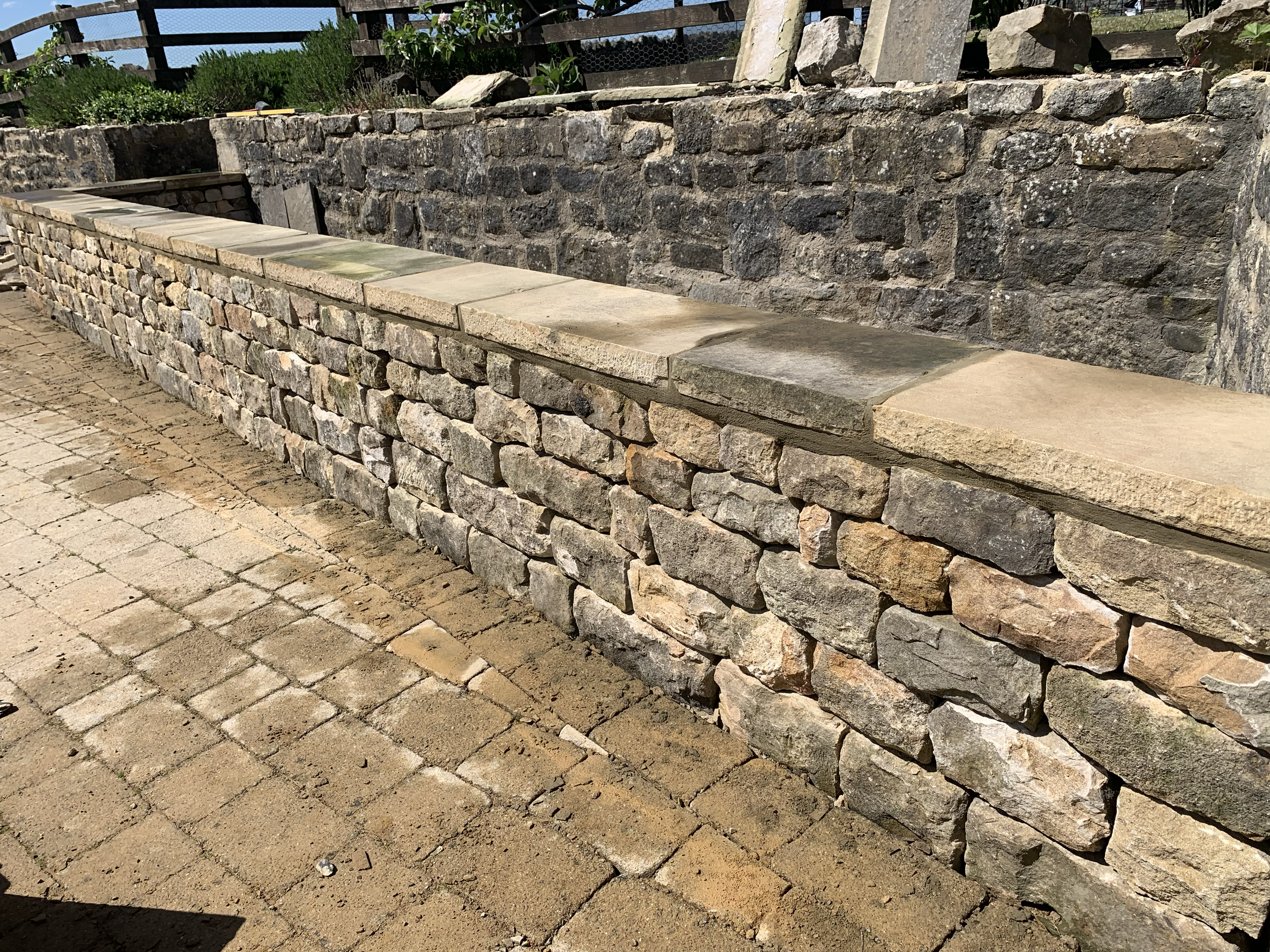 A dry stone retaining wall made of stacked, uneven stones with a smooth stone cap on top, situated on a paved brick sidewalk in bright sunlight.