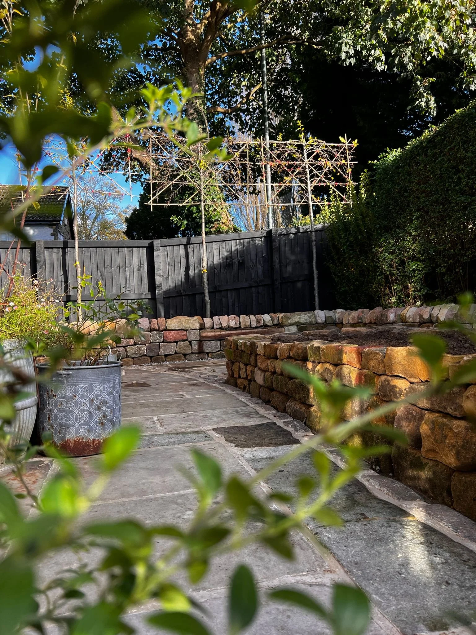A garden patio with stone paving, potted plants, a dark wooden fence, and a metal garden structure against the background of trees and bushes.