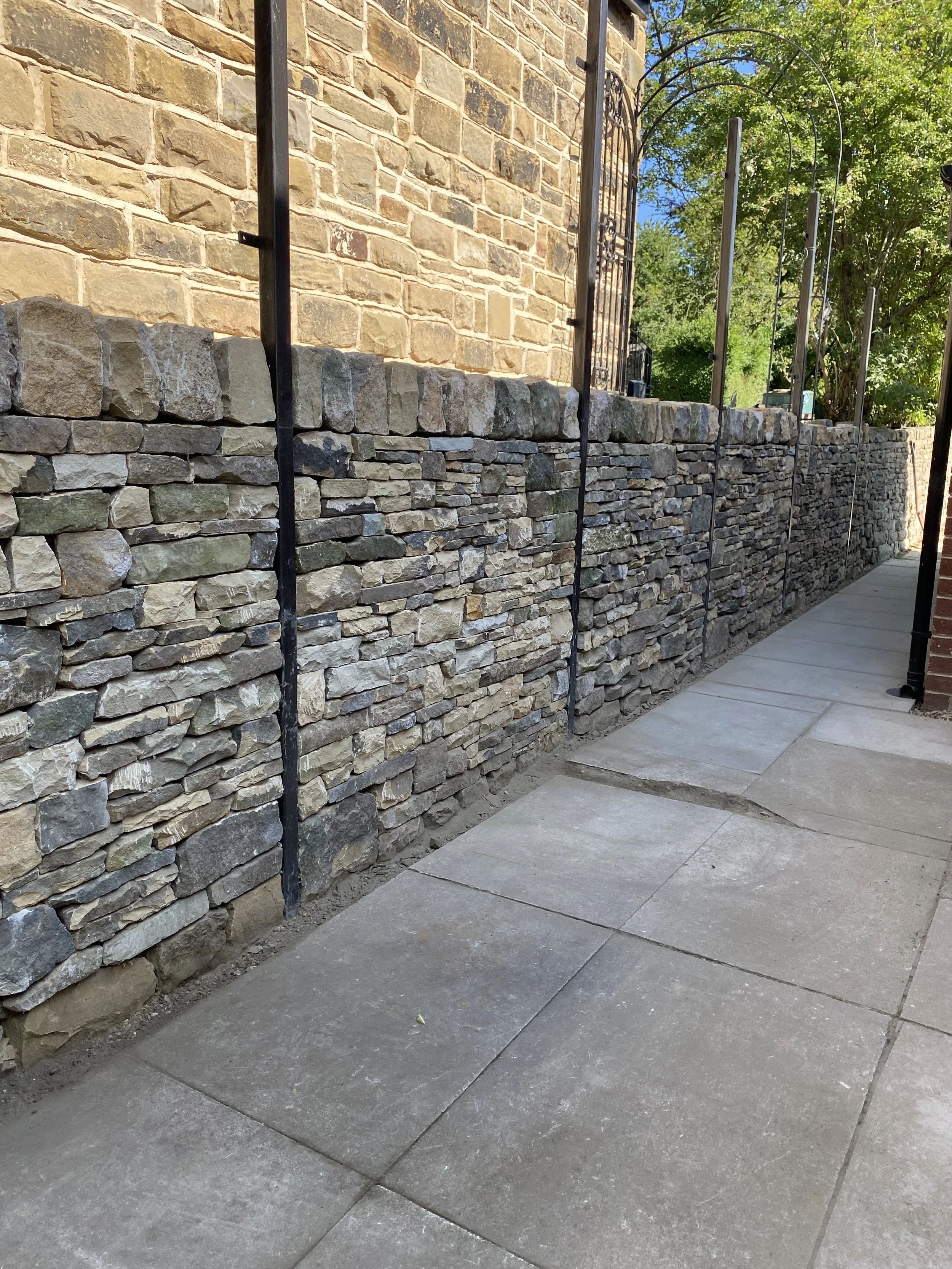Photo of a dry stone wall with a brick building behind it, along a concrete sidewalk with paving stones, green trees, and blue sky in the background.