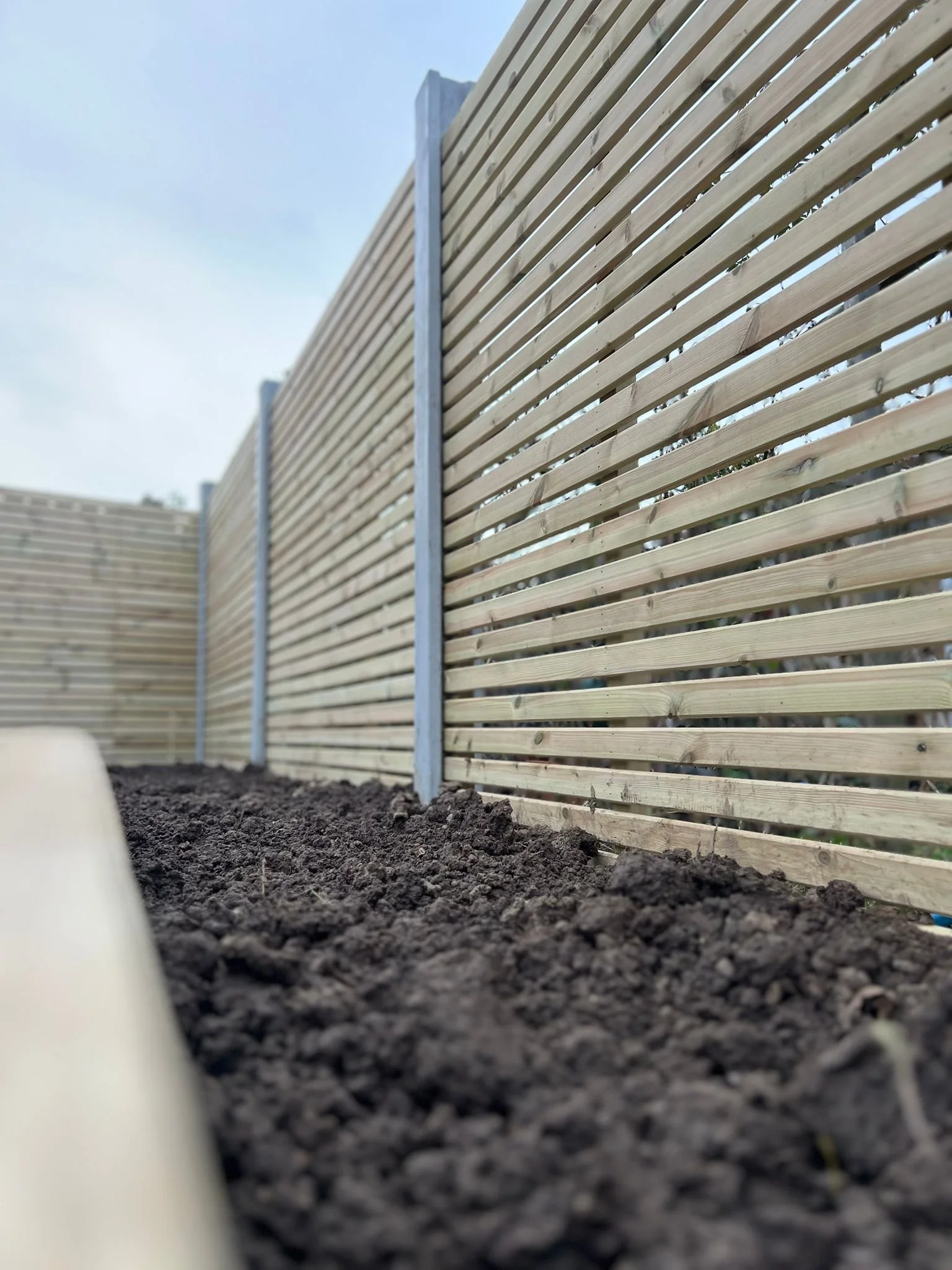 Wooden slat fence with metal posts on a bed of dark soil, under a cloudy sky.