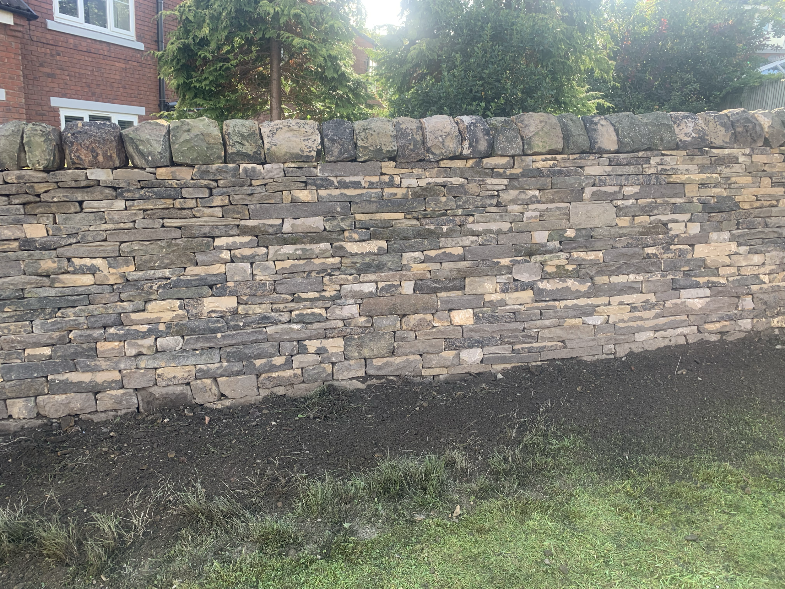 A dry stone brick wall with a row of rounded stones on top, situated in front of a house with trees and greenery in the background.