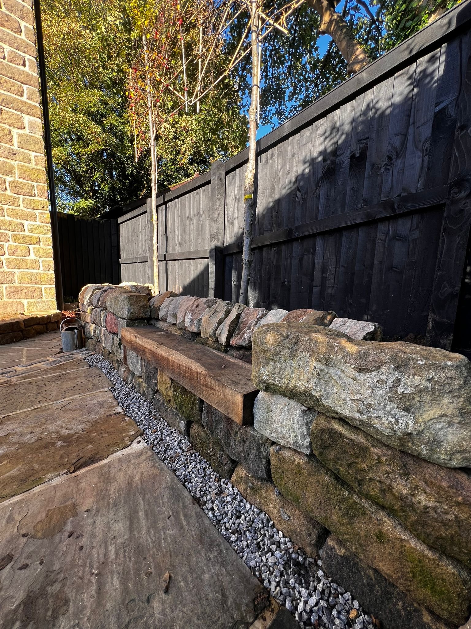 A dry stone wall with a wooden bench, small gravel bed, and two small trees against a black wooden fence, with sunlight and clear blue sky in the background.