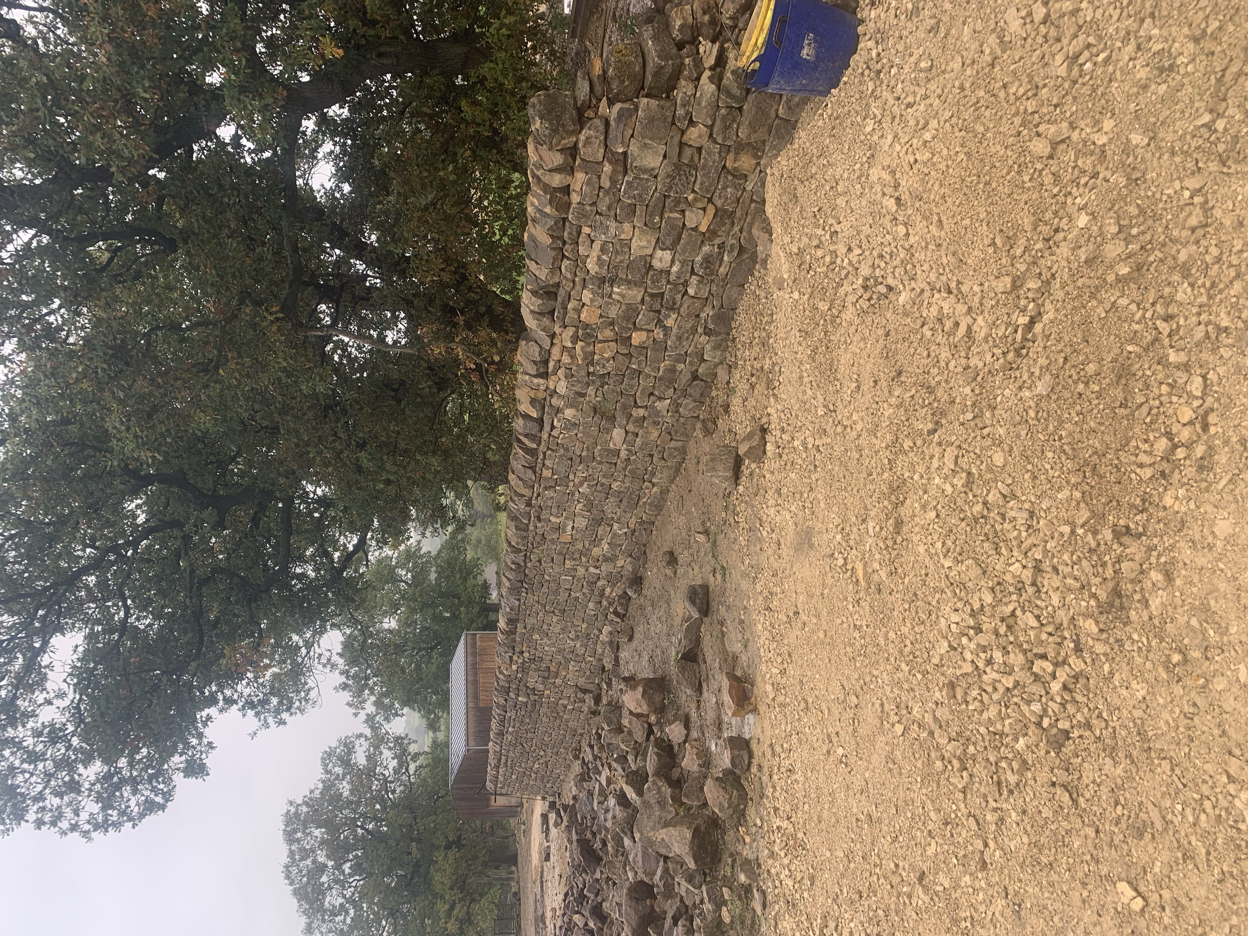 A dry stone wall with an uneven top edge, situated in a natural outdoor setting with trees and foggy sky in the background, and a blue plastic container on the ground nearby.