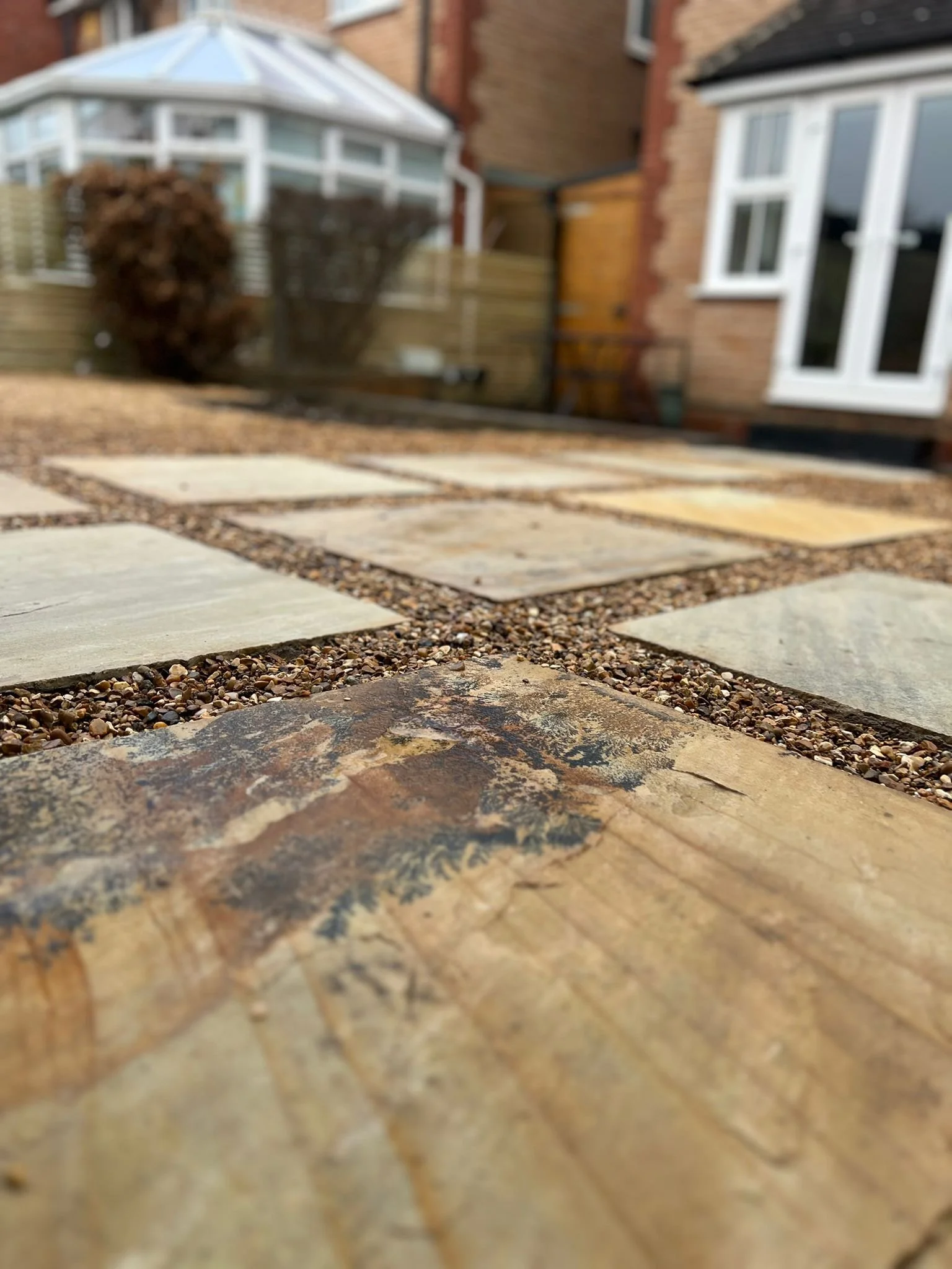 Close-up view of a stone and gravel patio in a backyard, with a house, window, fence, and garden shed in the background.