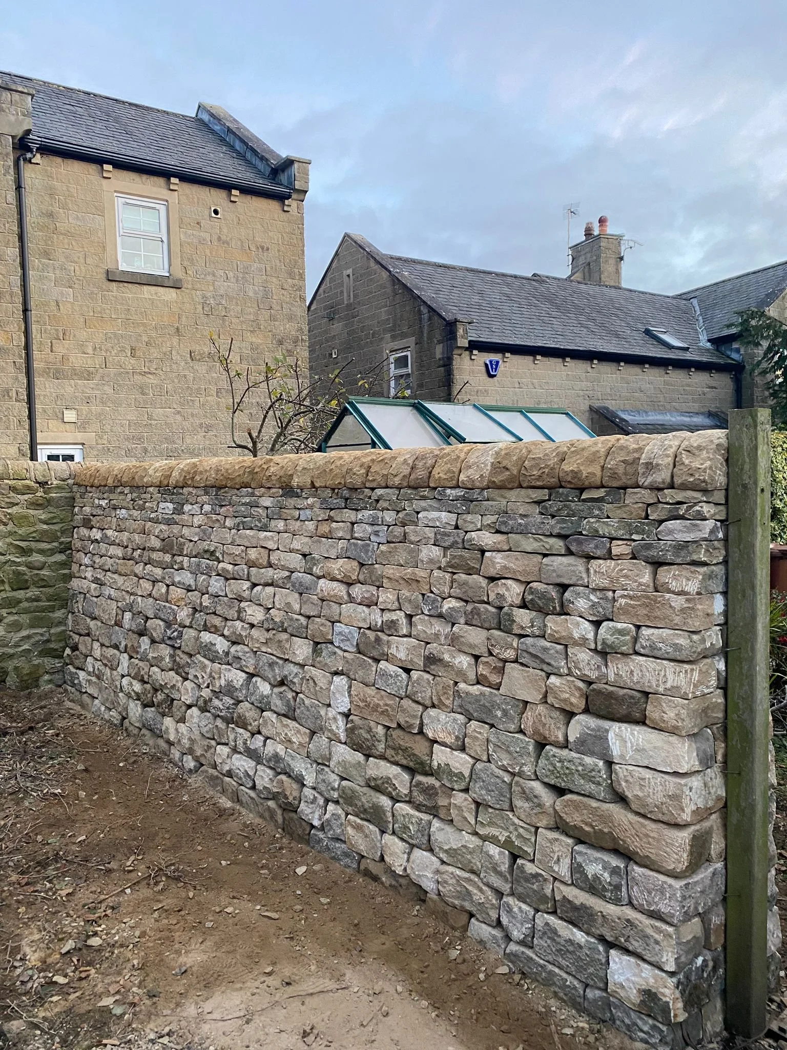 A dry stone wall in front of residential houses with gray roofs and chimney stacks, and a greenhouse with glass panels in the background under a cloudy sky.