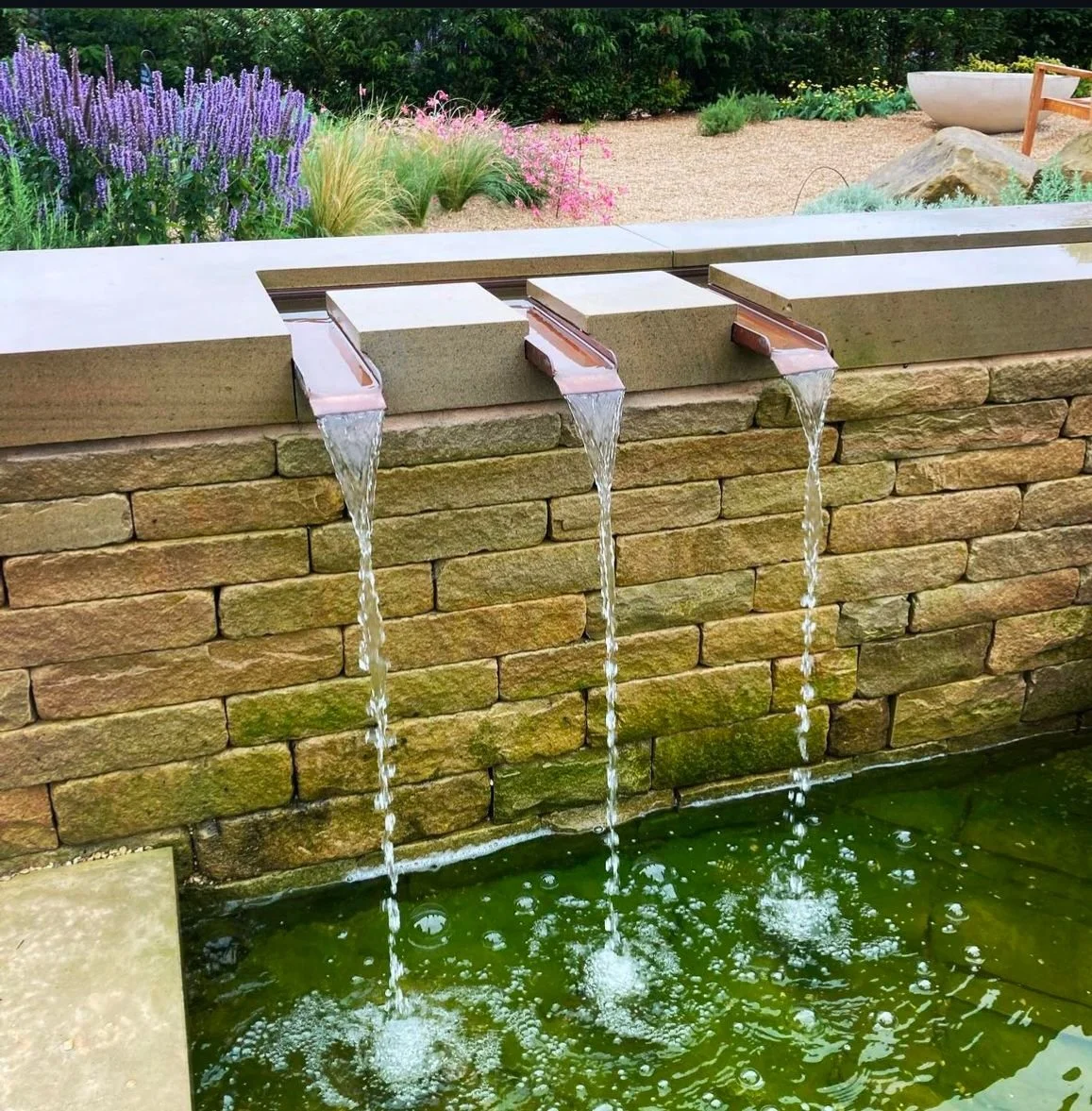 Water feature with three streams flowing from rectangular spouts over a dry stone wall into a pond below, with a garden background.