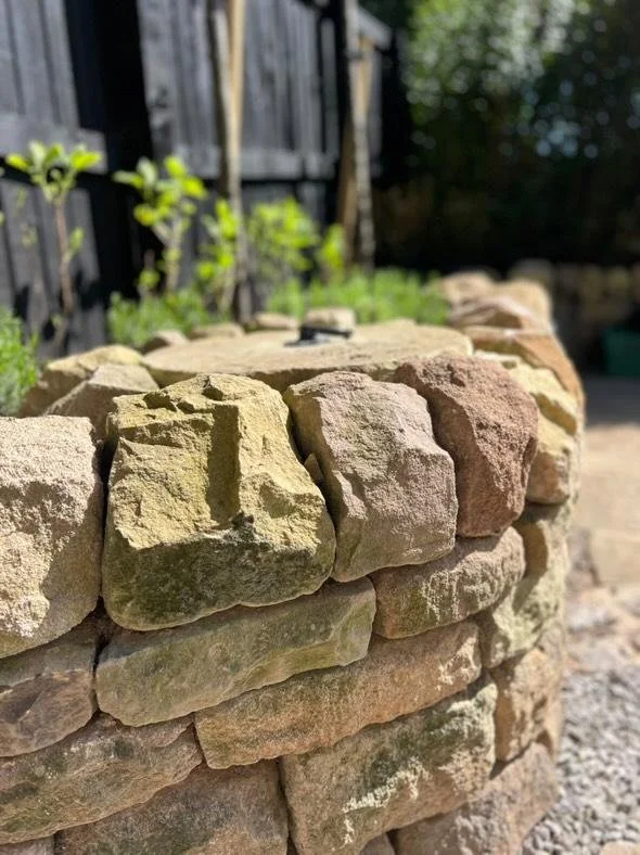 Close-up of a dry stone wall made from irregularly shaped stones with a garden fence and greenery in the background.