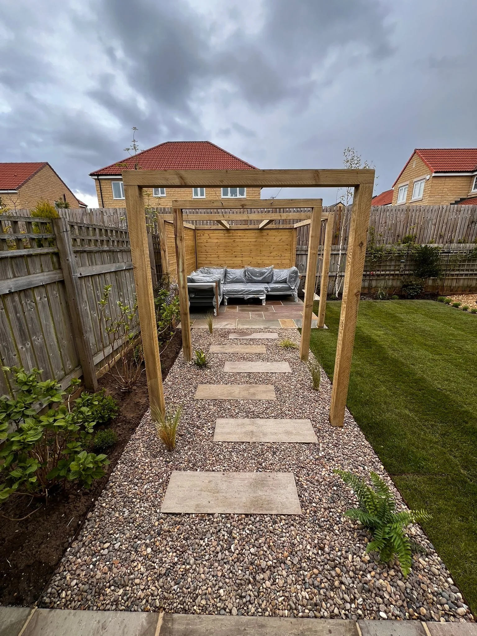 A backyard with a gravel pathway, wooden pergola framework, and a seating area with a plastic-covered sofa, surrounded by a wooden fence and bordered by grass and plants, under cloudy sky.