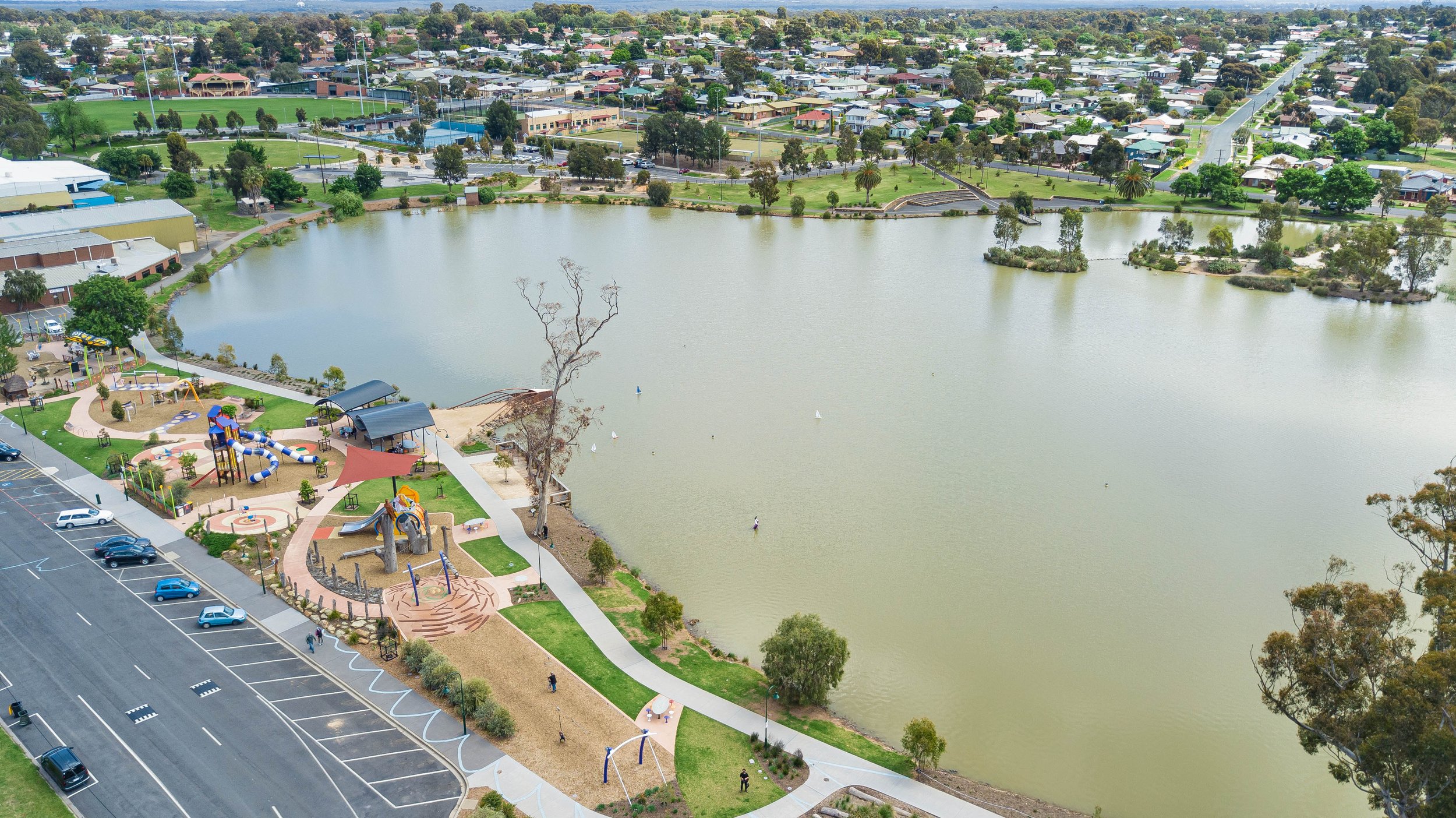 Aerial view of a lakeside park with walking paths, playground equipment, and picnic areas. The lake is surrounded by residential neighborhoods and green spaces. Several cars are parked along a nearby road.