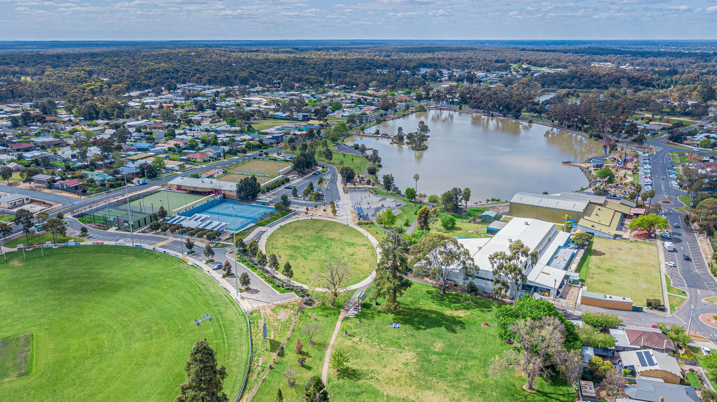Aerial view of a suburban area with a lake, sports fields, tennis courts, houses, and surrounding greenery.
