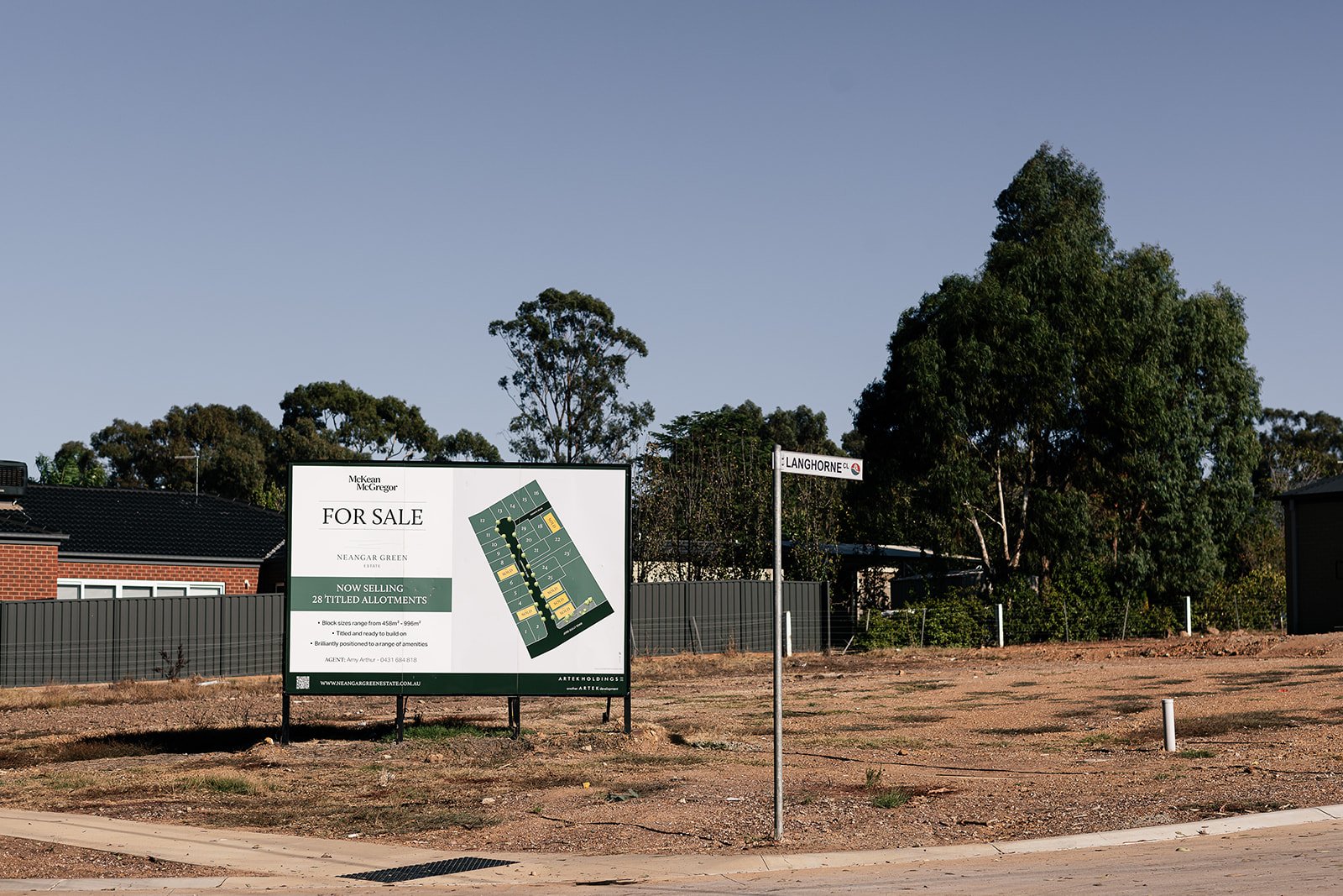 Vacant lot with a "For Sale" sign advertising 28 titled allotments, situated at the corner of Langhorne Street, surrounded by trees and a fence, with a neighboring building visible.