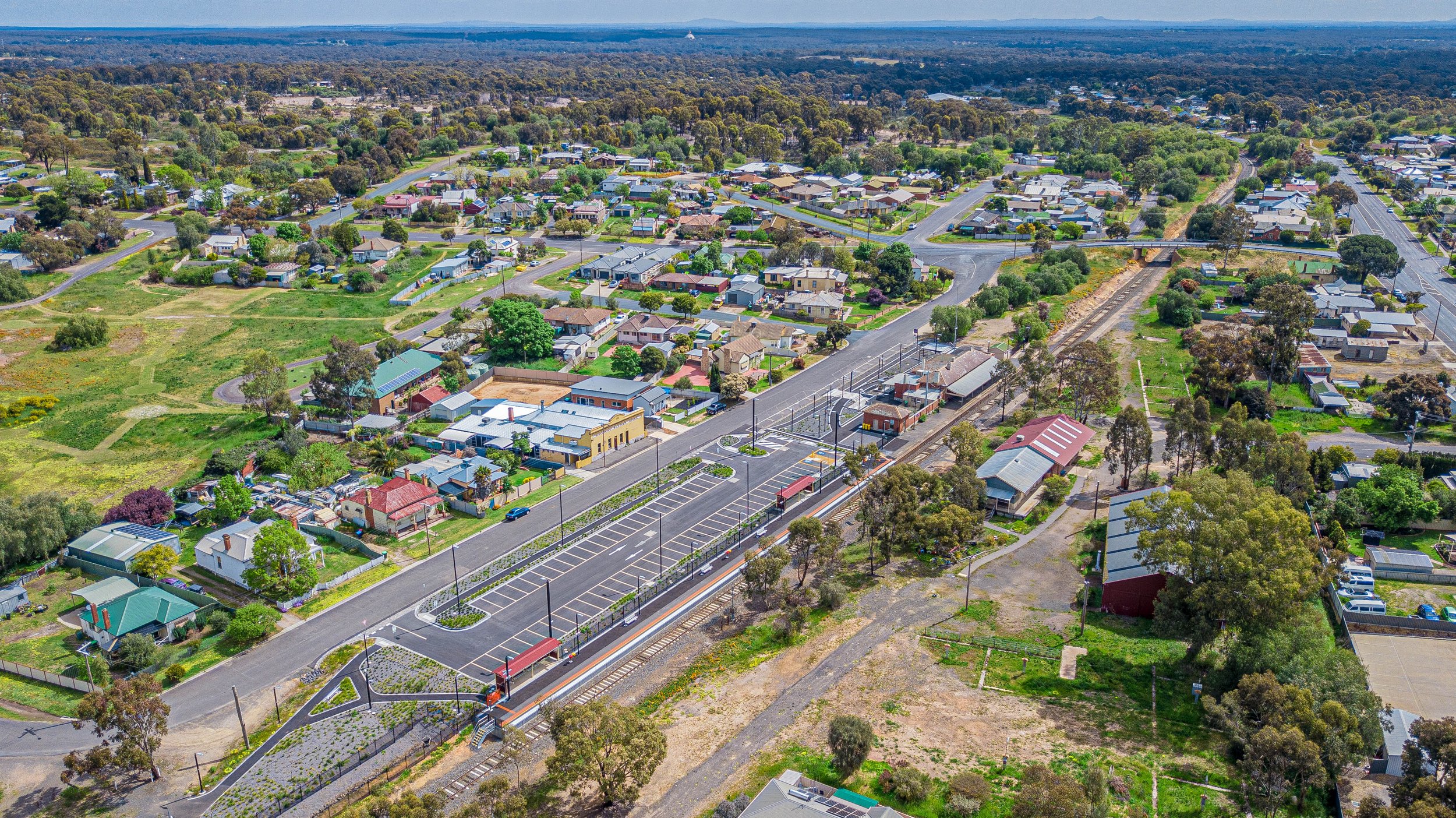Aerial view of a small town with scattered houses, roads, and green spaces. A railway line runs through the area, with a train station and a parked train visible. The landscape shows a mix of trees, grassy fields, and urban development.