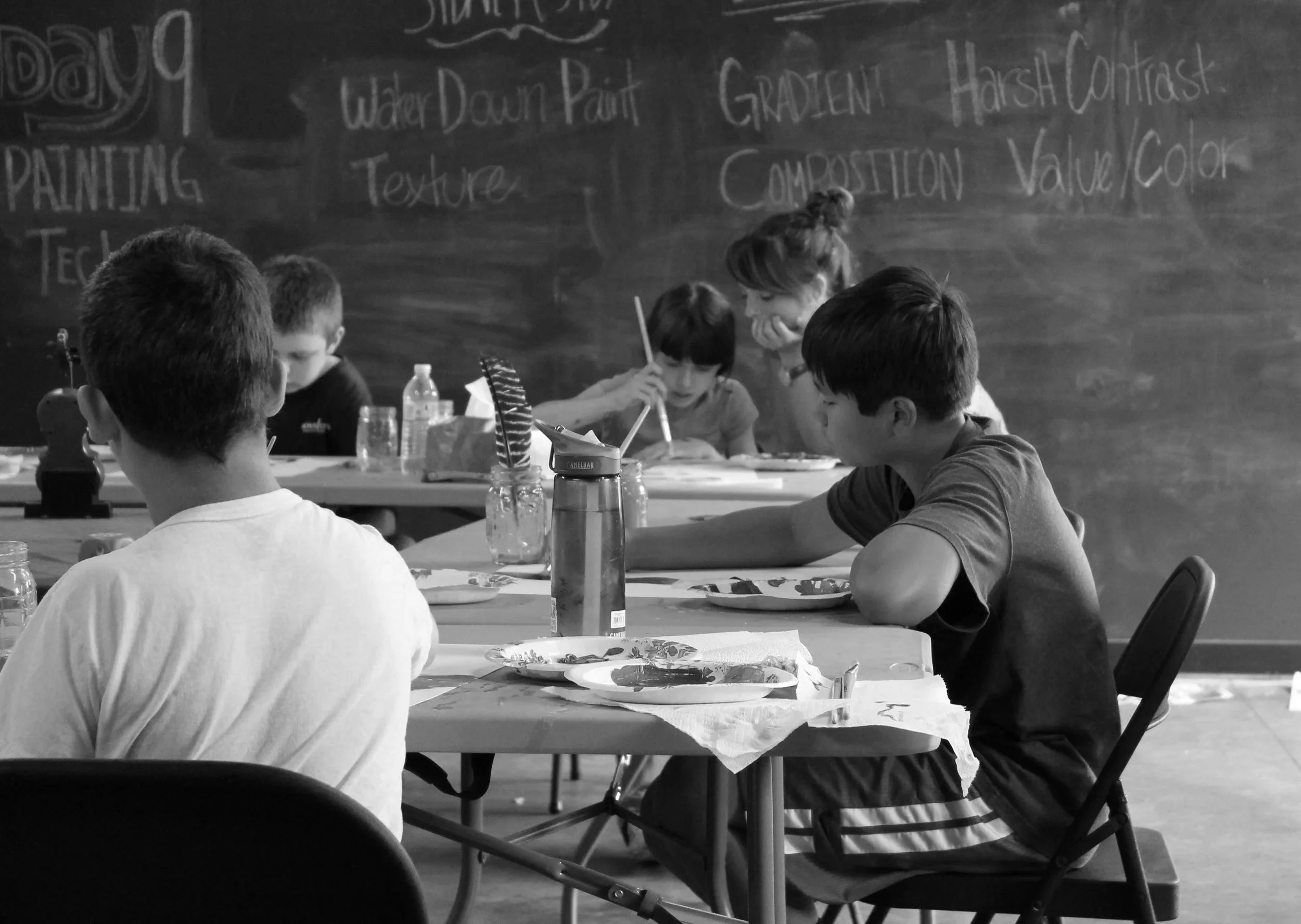 Children sitting at tables participating in an art class, with a chalkboard in the background featuring notes on watercolor painting techniques.