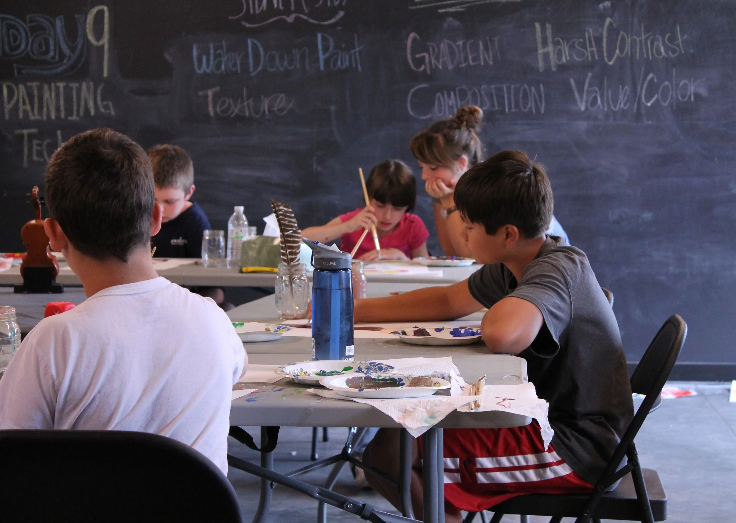 Children engaged in painting activities at a classroom table with art supplies, a teacher assisting a girl, and a blackboard with art lesson topics written on it.